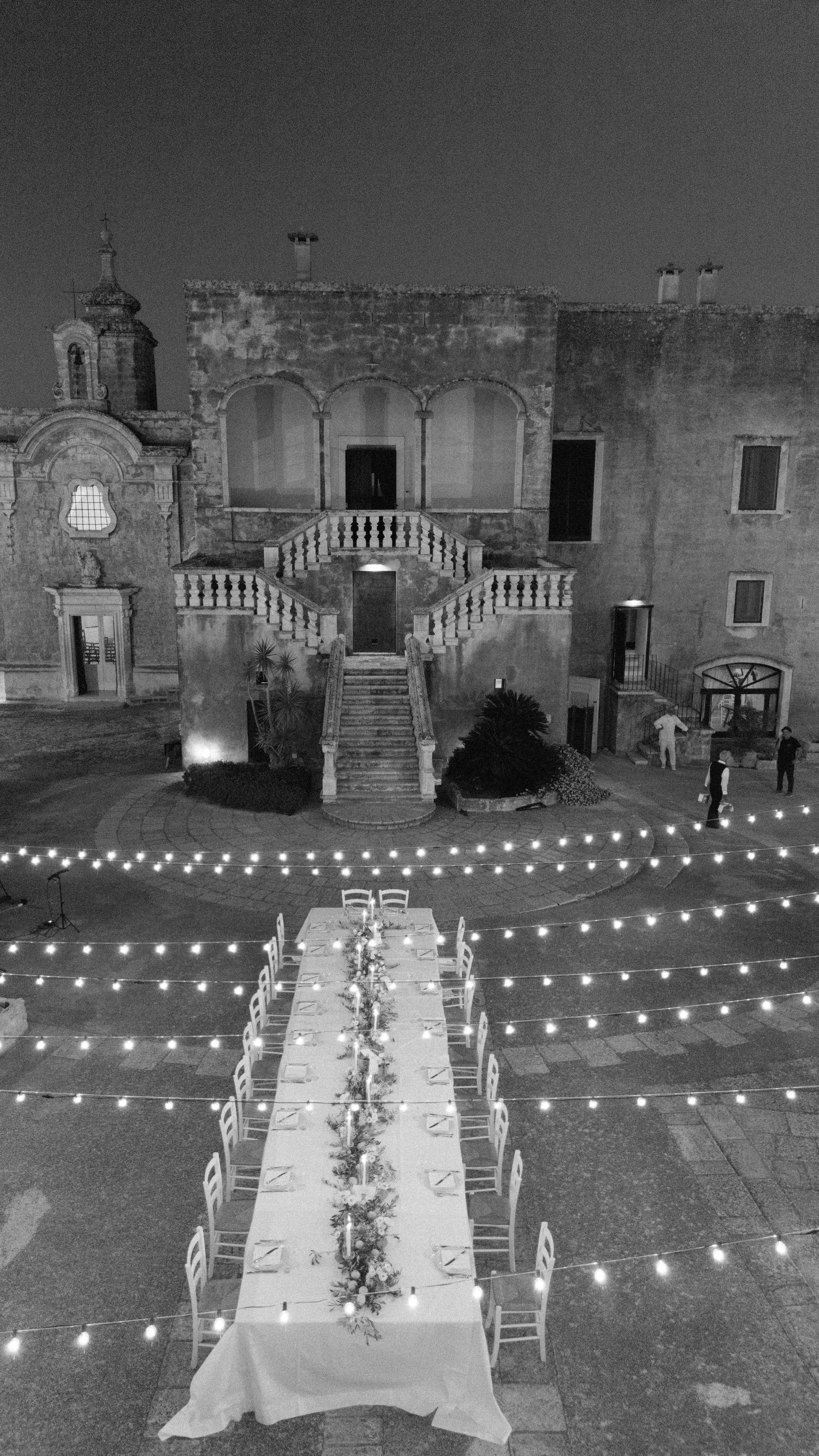 A long dining table set under string lights leads to an elegant stone staircase of a historic Mediterranean villa at night.