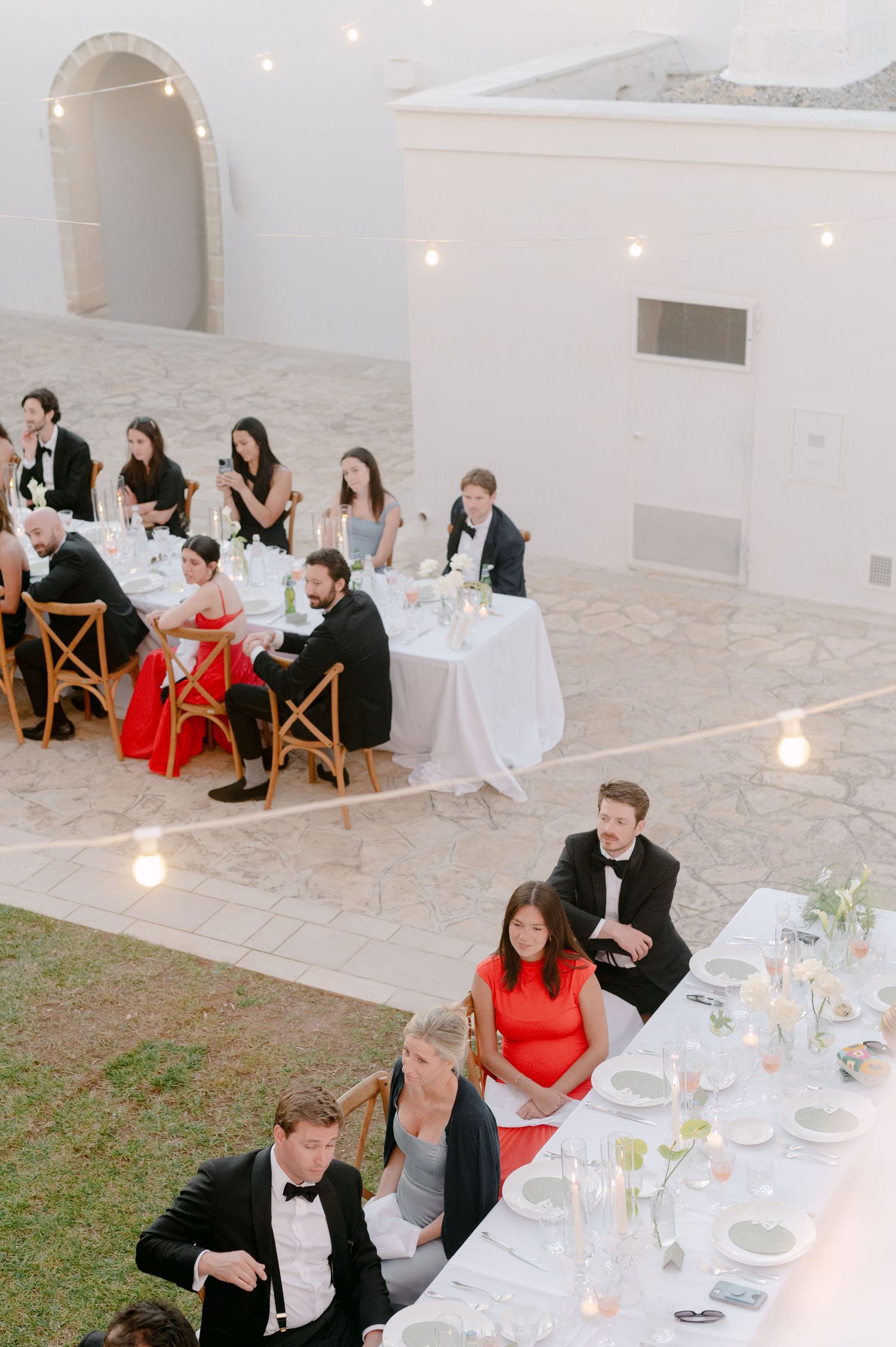 Elegant outdoor wedding reception with guests in formal attire seated at long white tables, adorned with candles and floral centerpieces.