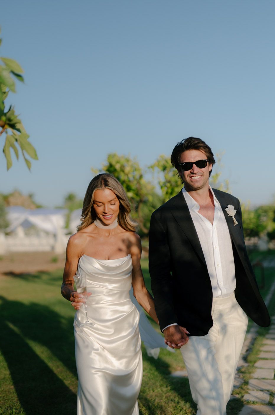 Couple in white and black formal attire walking along a sunny outdoor path.