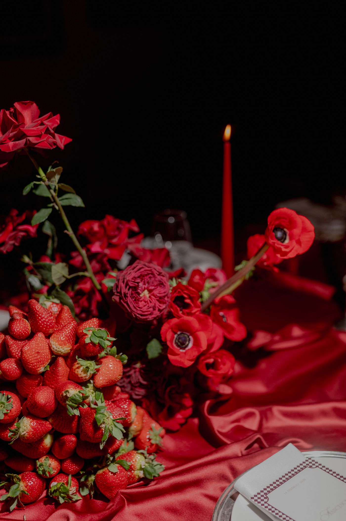 Romantic red roses and fresh strawberries arranged on silky red fabric with glowing candles creating an intimate atmosphere.