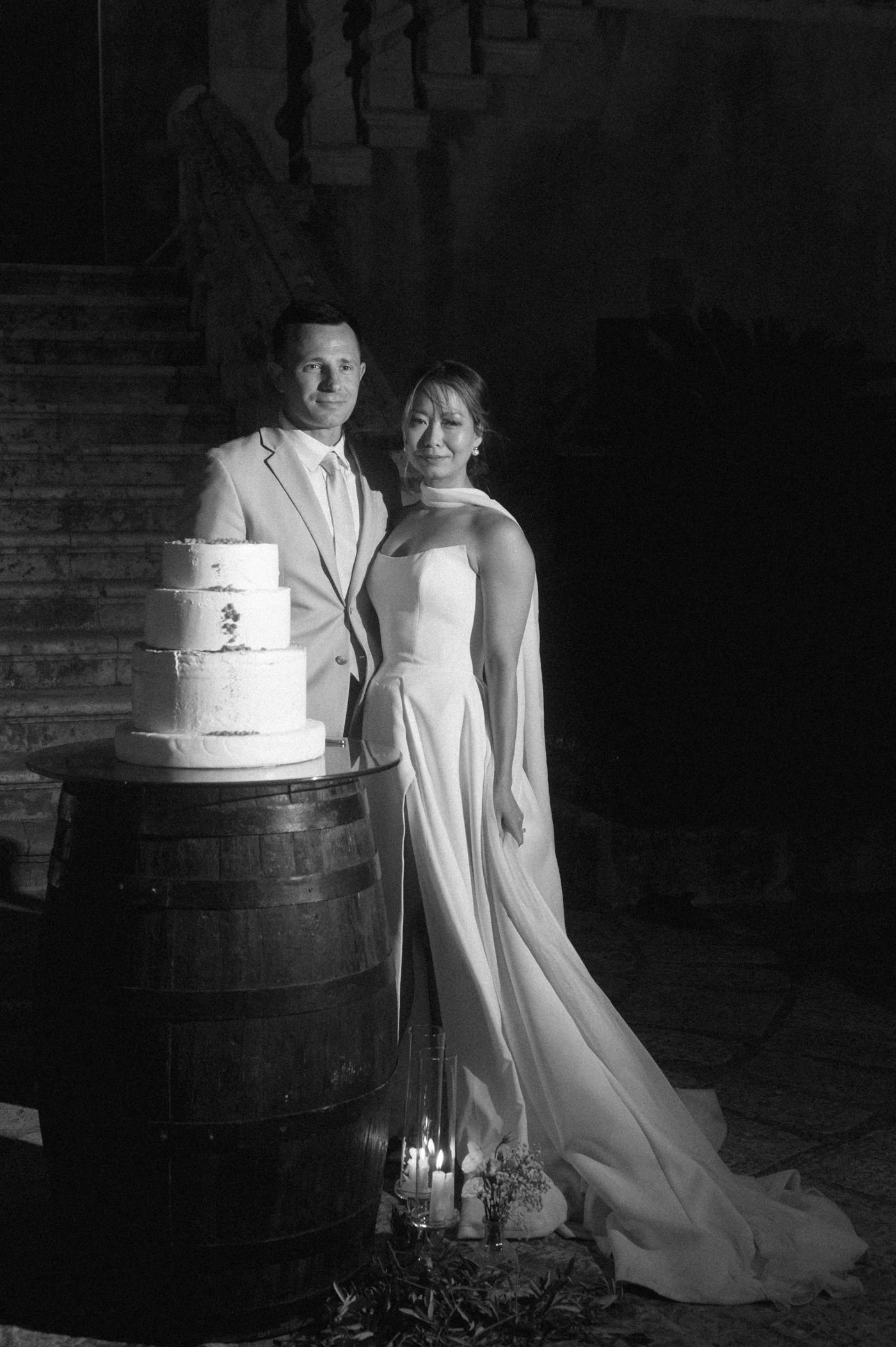 A dramatic black and white wedding photo series showing a bride and cake displayed on a rustic wooden barrel.