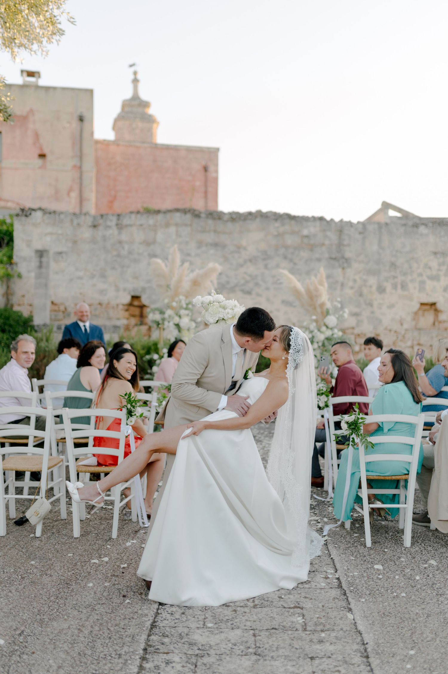 Wedding guests mingle at an outdoor reception with white chairs and stone walls at a rustic venue during sunset.