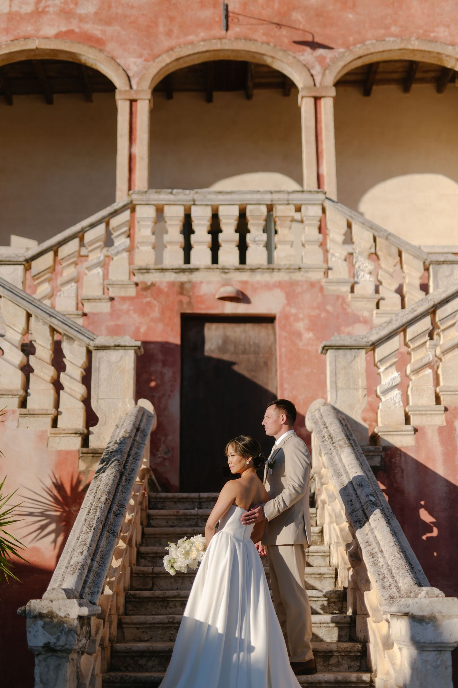 A couple stands on an elegant pink stone staircase with ornate balustrades at a historic Mediterranean-style villa.