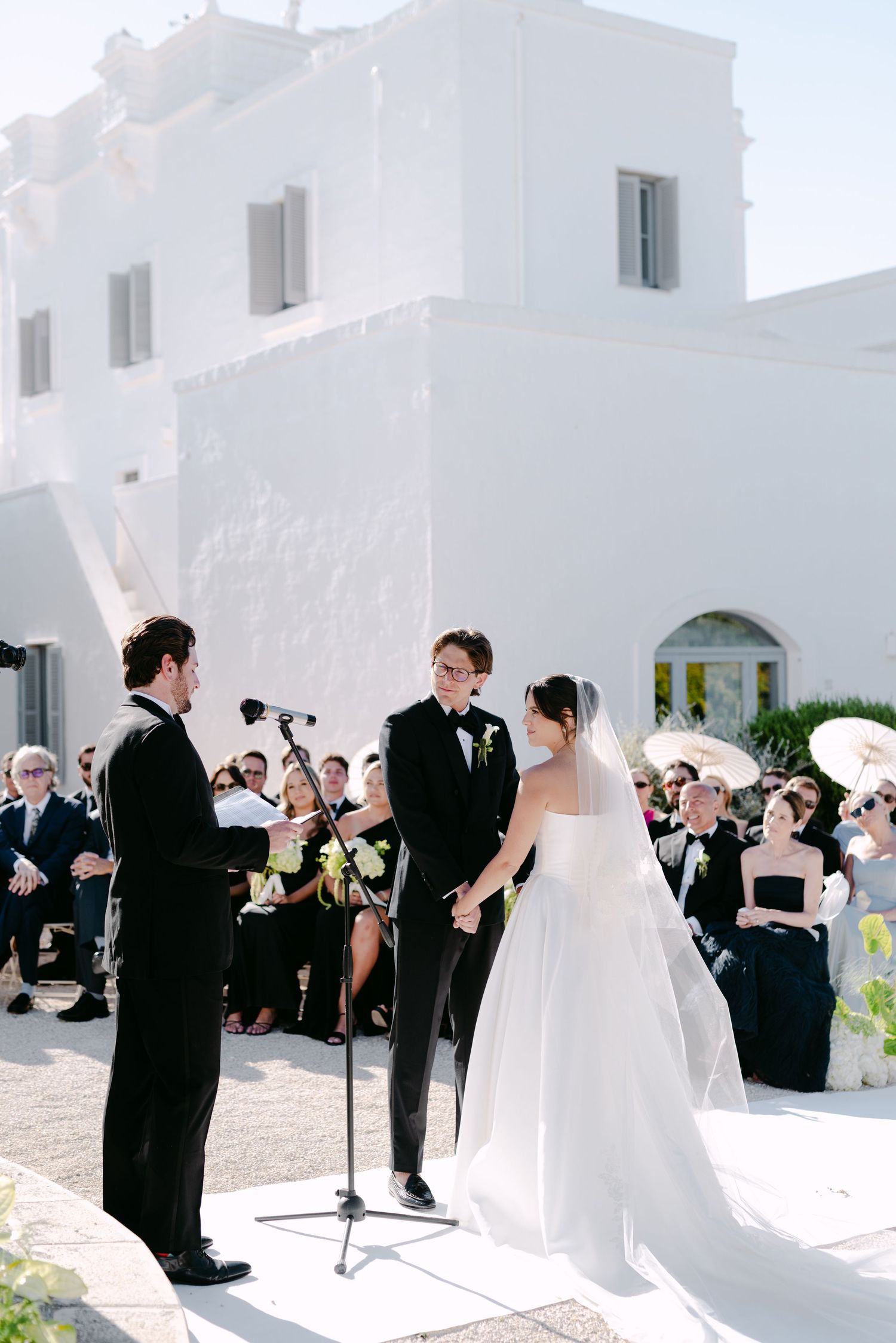 Bride and groom exchange vows at an outdoor wedding ceremony beside a white Mediterranean-style building.