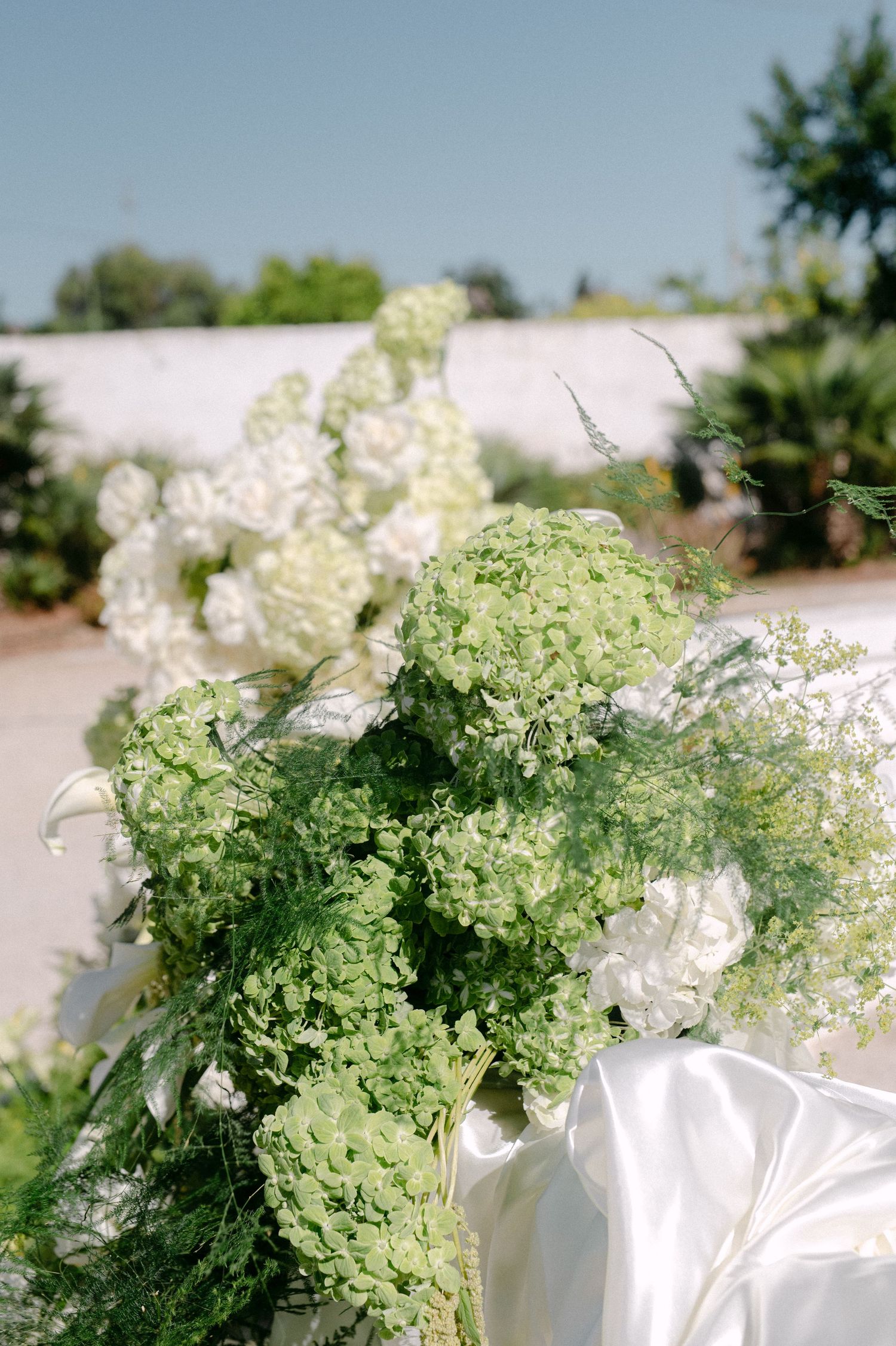 Lush green and white hydrangea wedding floral arrangement with soft white satin ribbon outdoors.