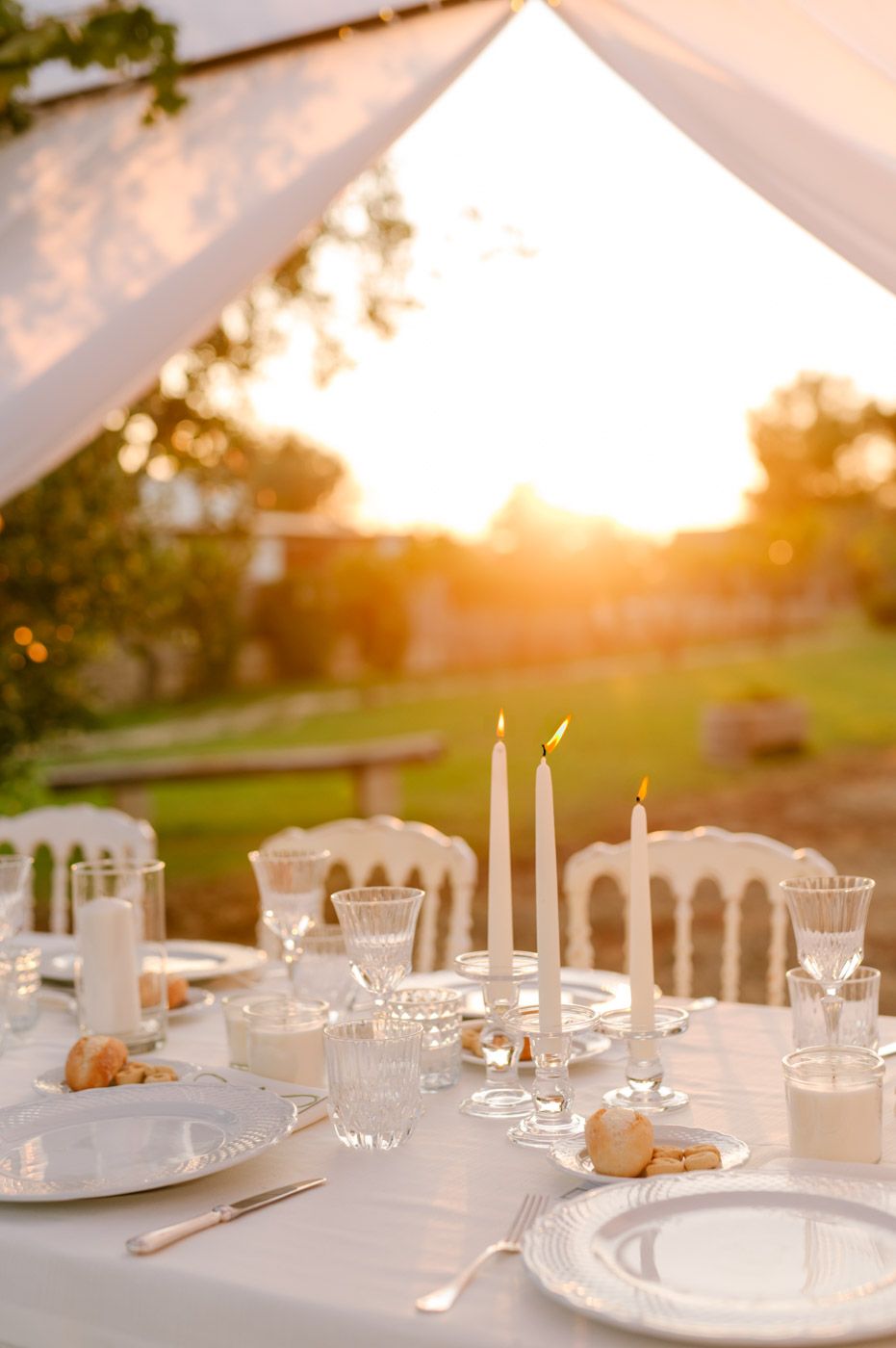 Elegant outdoor table settings with white candles and place settings glowing in golden sunset light at a formal dinner event.
