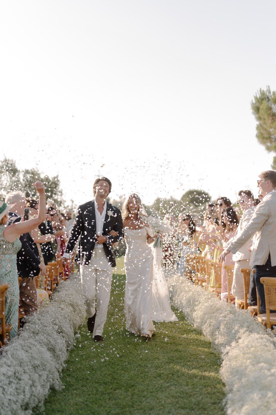 Wedding guests throw white flower petals during an outdoor celebration on a grassy hillside.