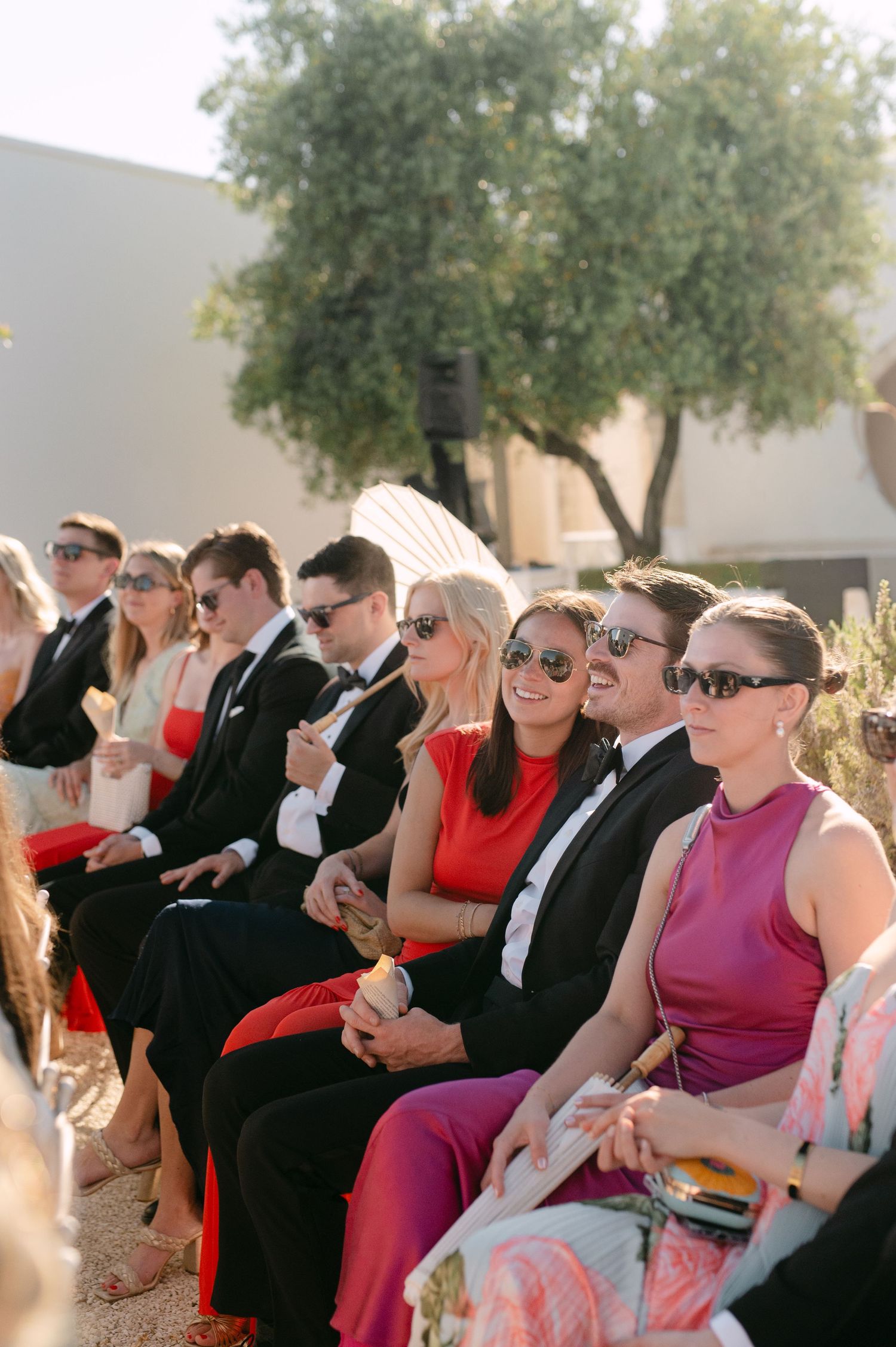 Wedding guests in formal attire and sunglasses seated outdoors, smiling during a sunny ceremony.