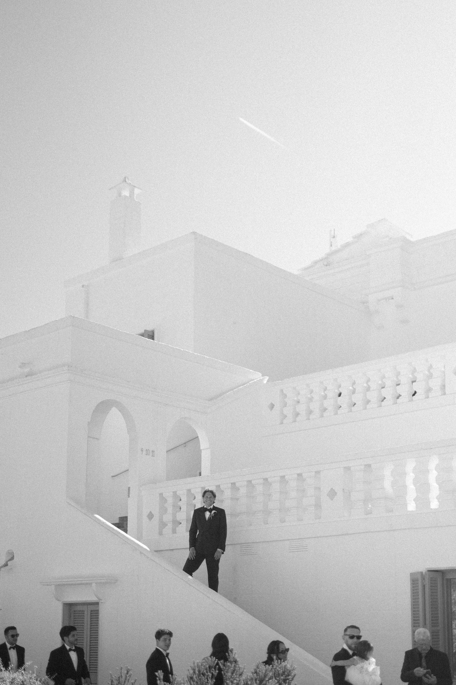 Black and white wedding photo of groom descending white Mediterranean villa stairs with guests gathered below.