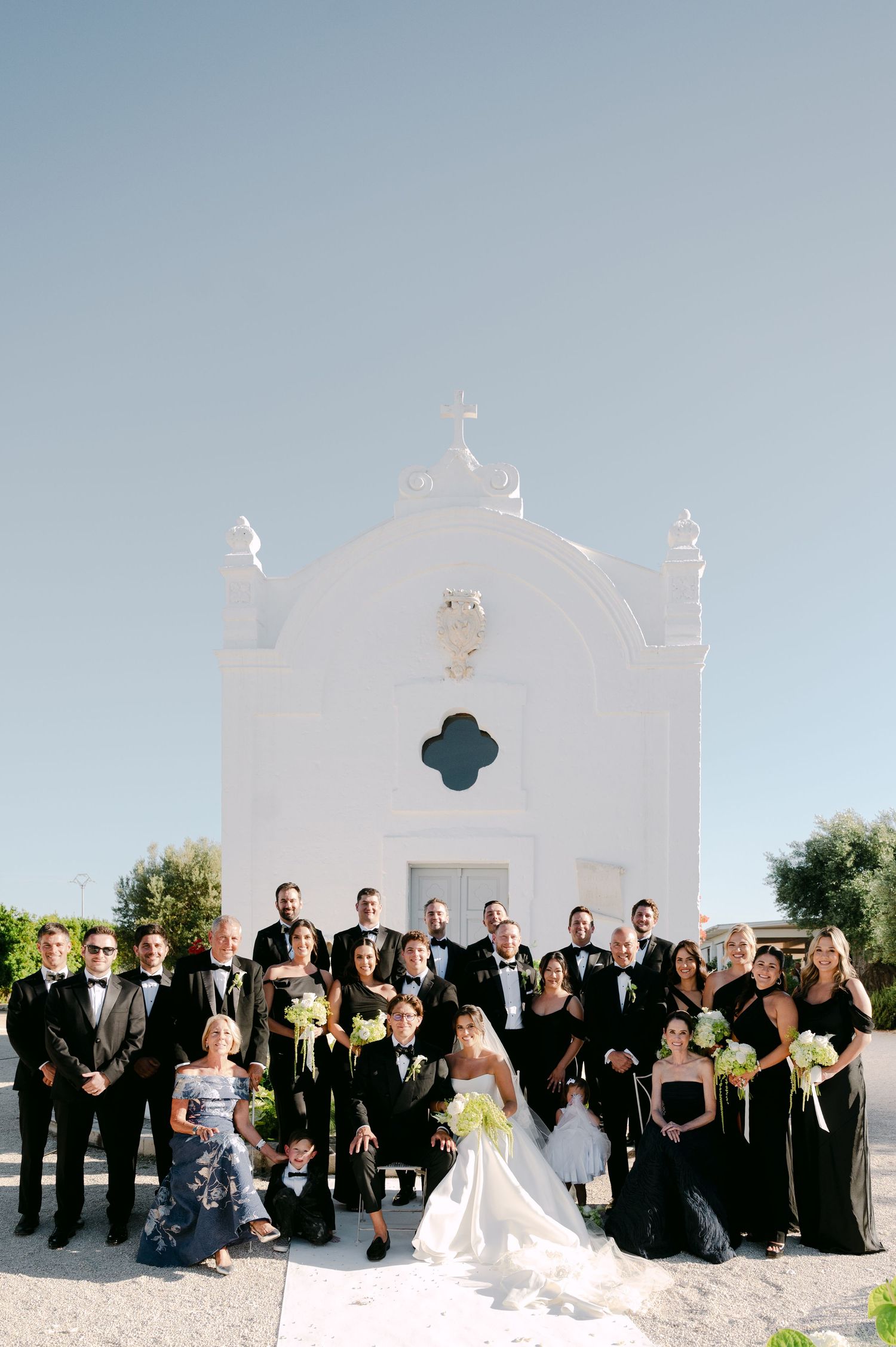 Wedding party in black attire poses before a white chapel with a cross under a clear blue sky.