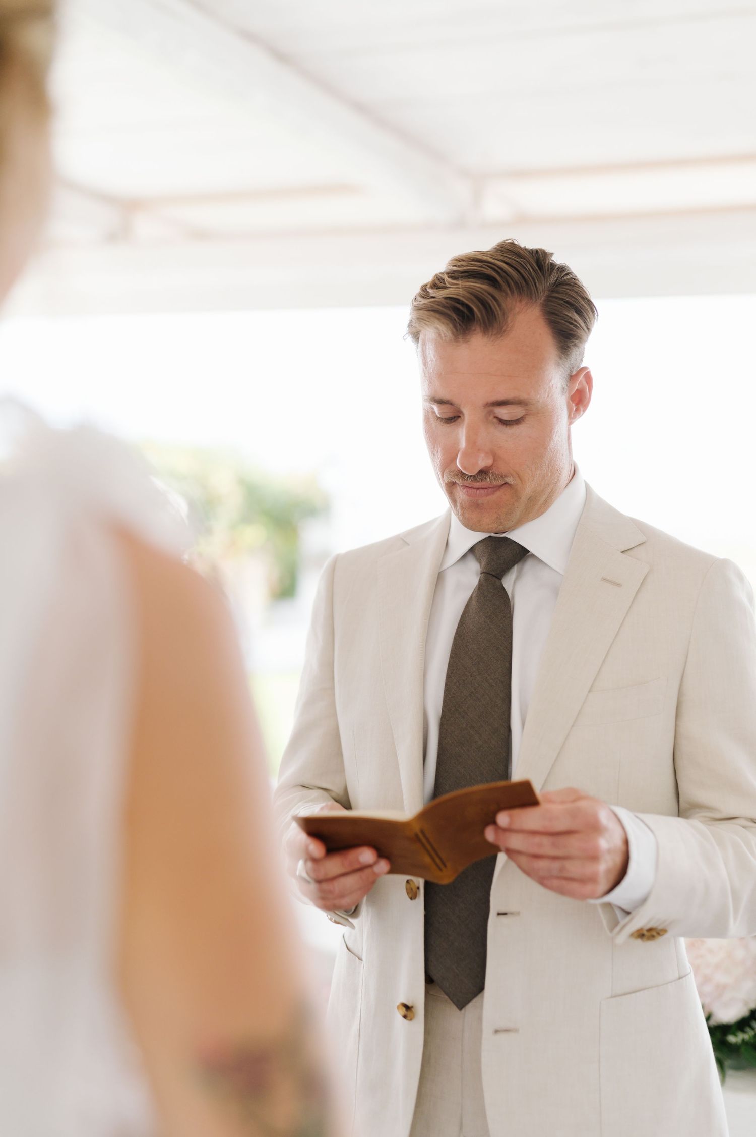 A person in a white suit reads from a small book in a sunlit room.