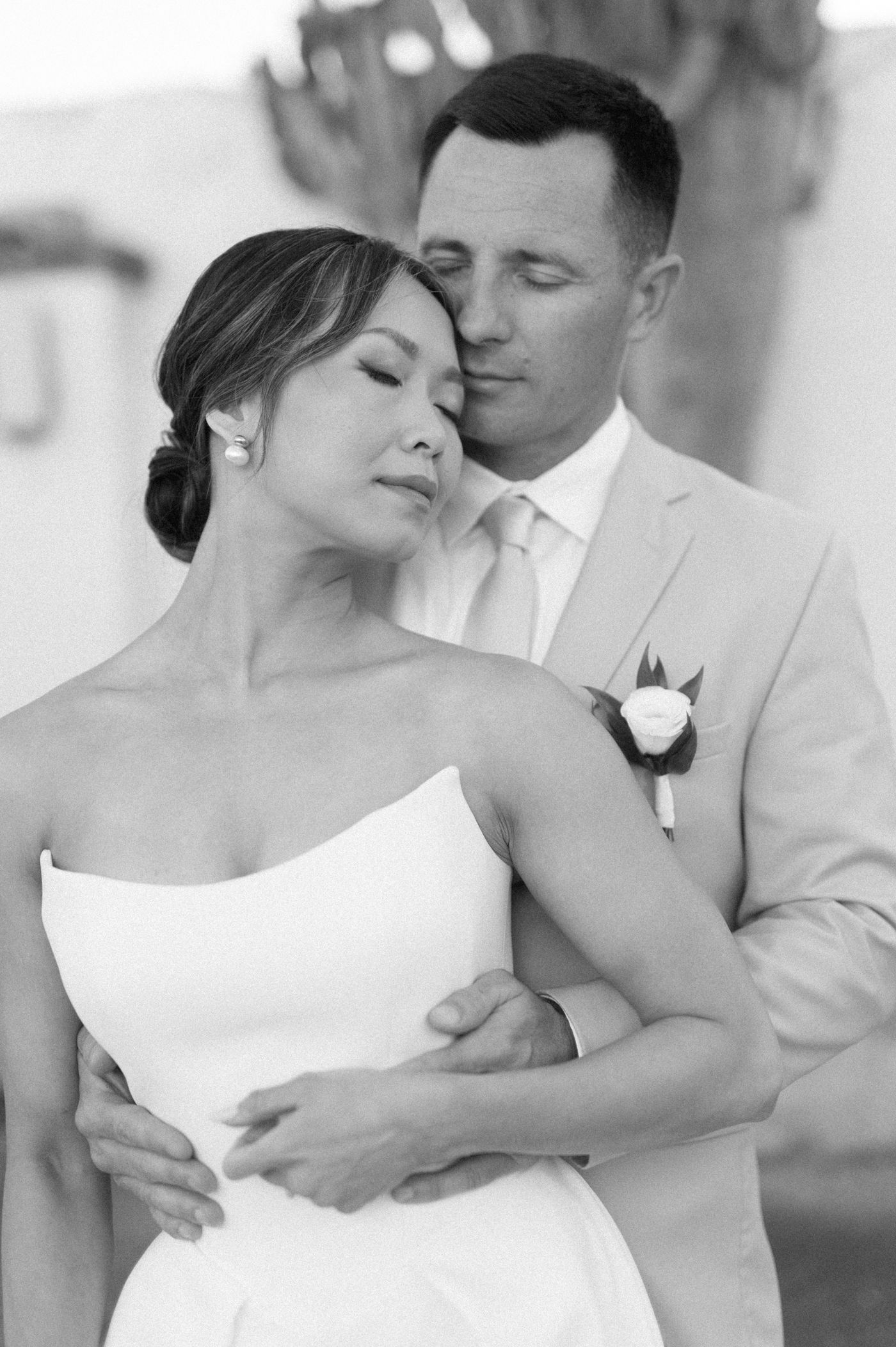 Intimate black and white wedding photo showing newlyweds in close embrace, bride in white dress and groom wearing boutonniere.