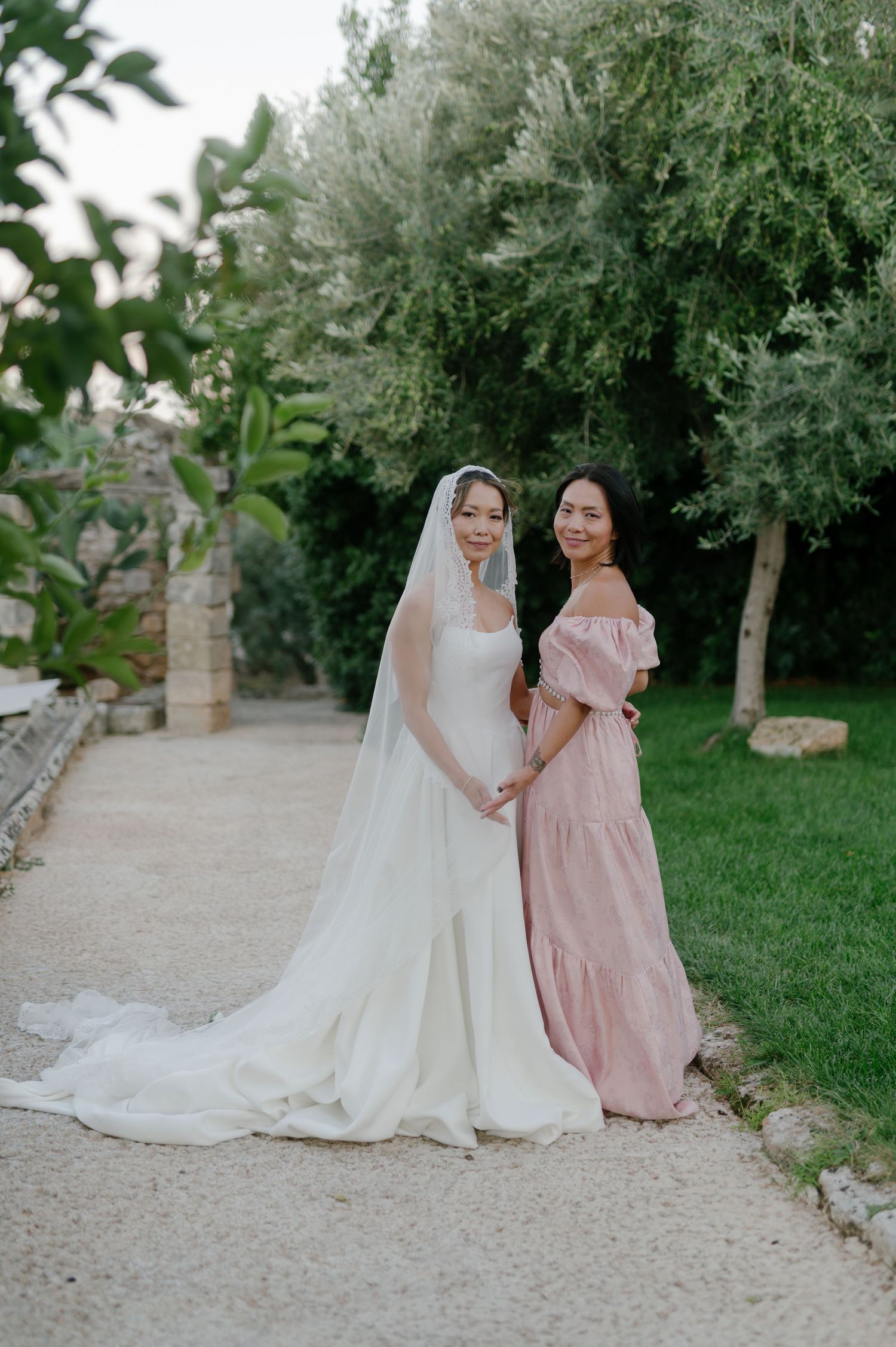 Two figures in elegant dresses, one in white bridal gown and one in blush pink, pose together in a garden setting.