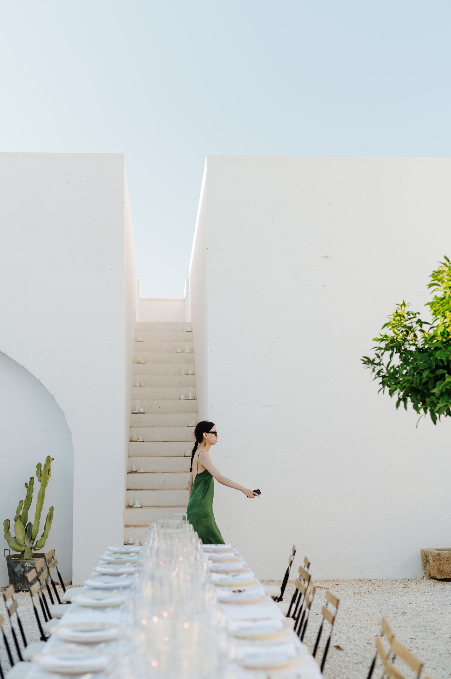 A minimalist outdoor dining setup with white walls, wooden chairs and a long table set for a special occasion.