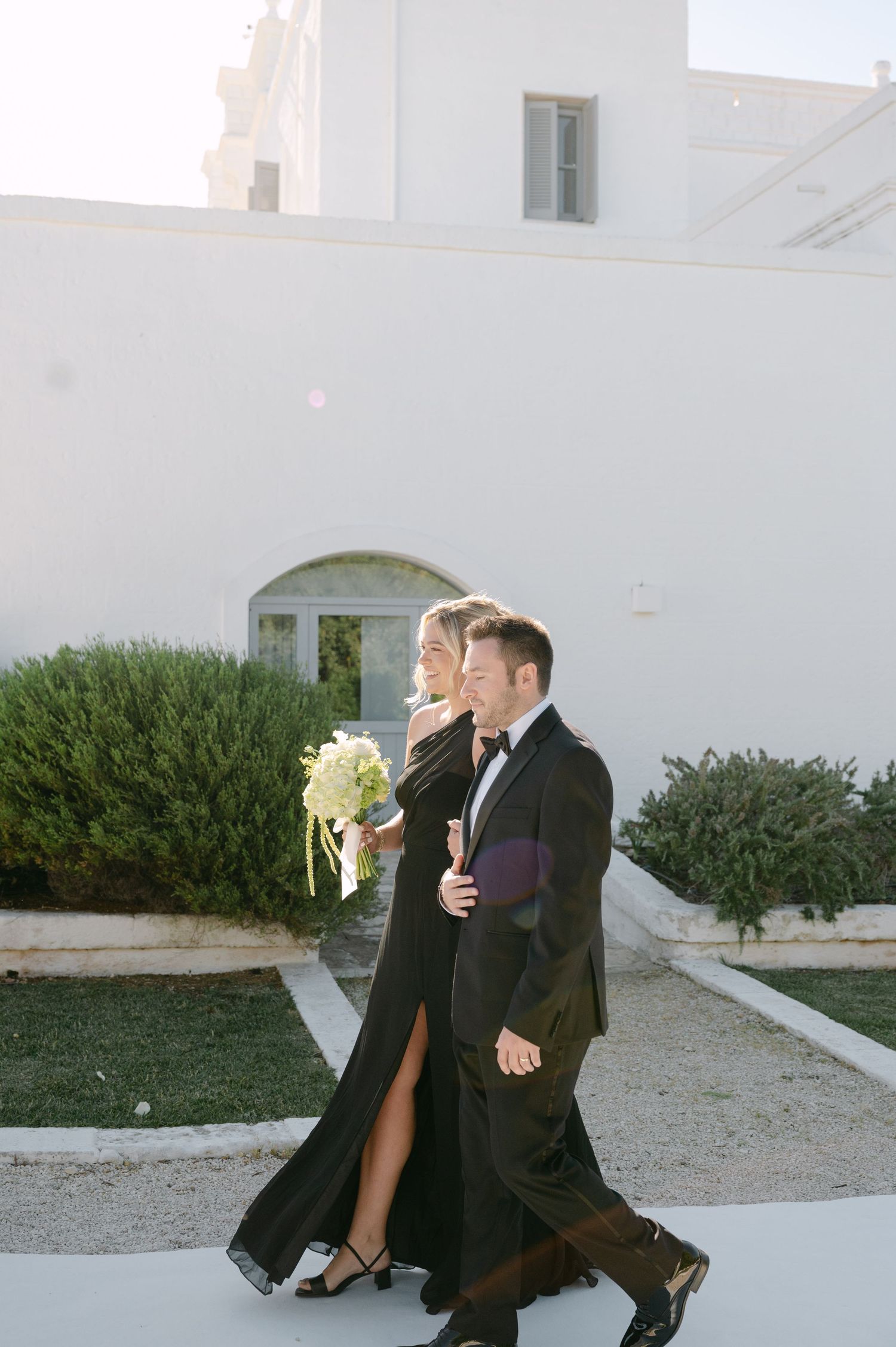 Bride in black gown holding bouquet walks with groom in tuxedo outside white Mediterranean venue.
