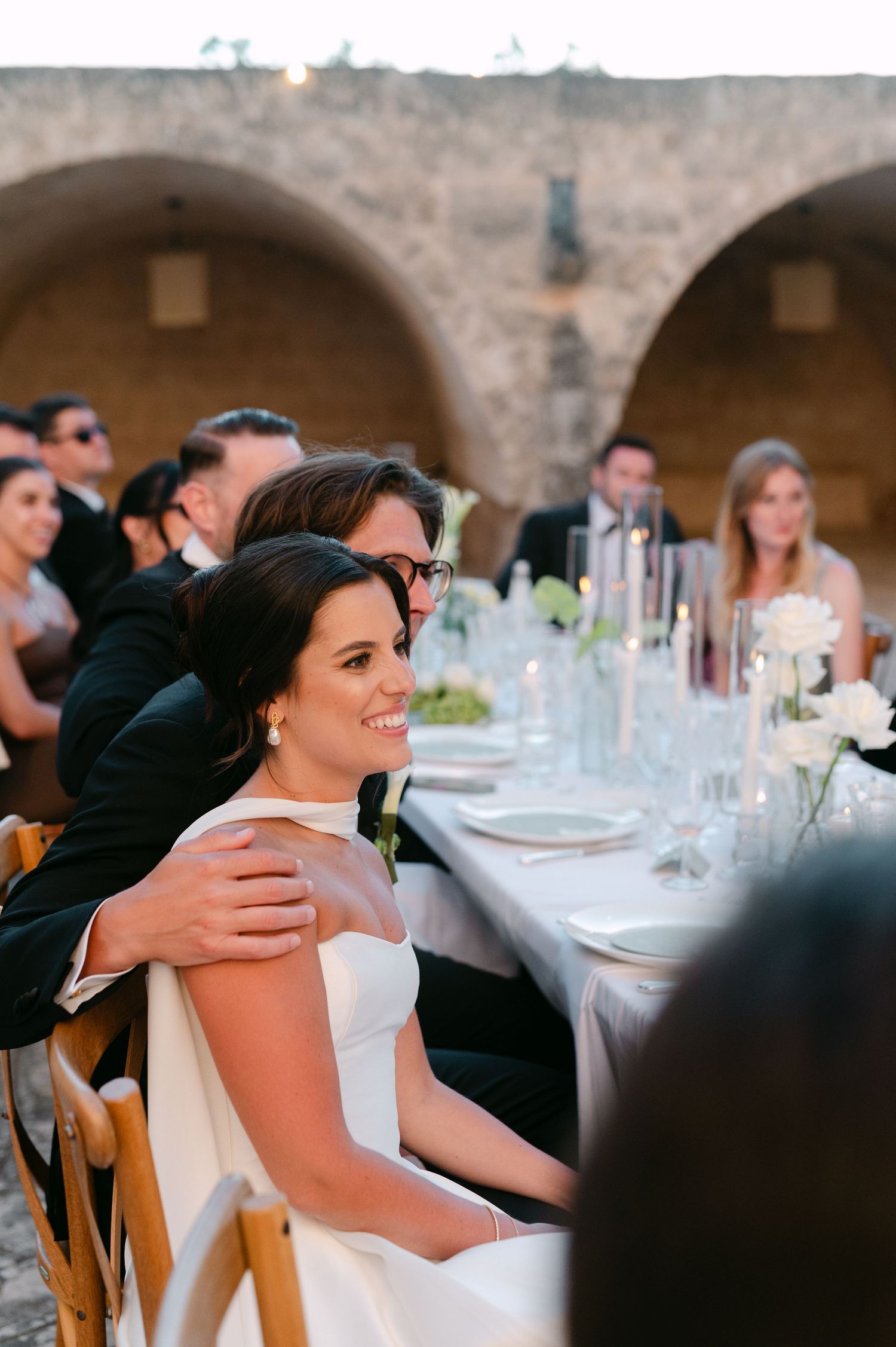 Bride in white gown smiles at outdoor wedding reception dinner with stone arch backdrop and elegant table setting.