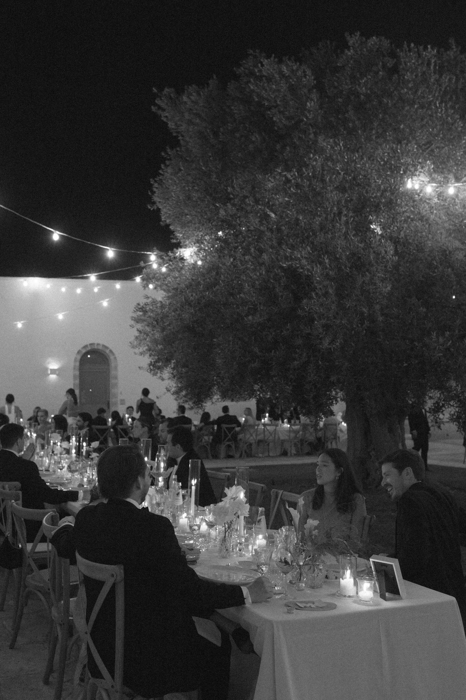 Black and white outdoor wedding reception at night with string lights, candles, and guests dining under a large tree.