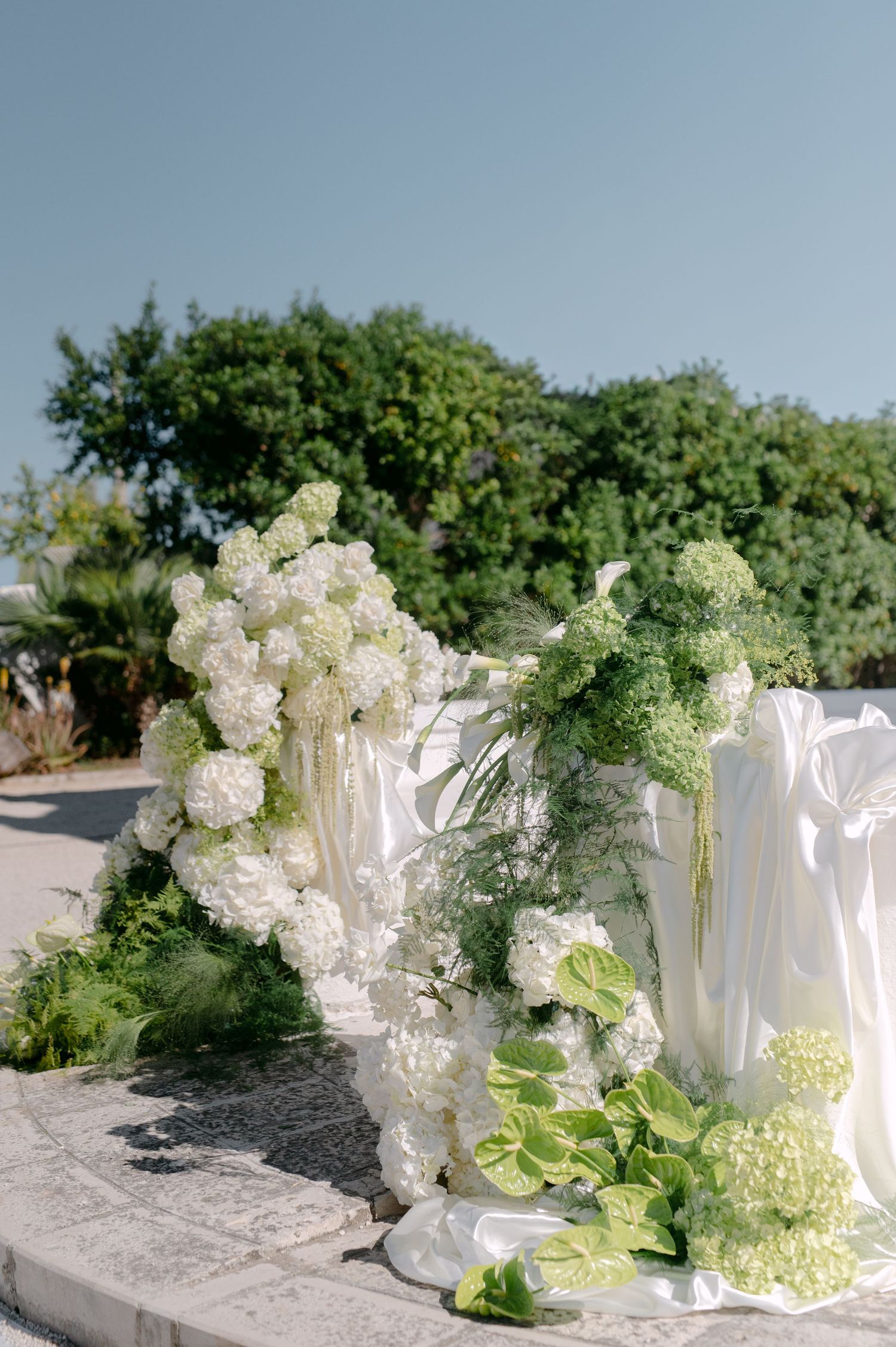 Elegant outdoor wedding ceremony decor with white hydrangeas, green foliage, and flowing white fabric against a clear blue sky.