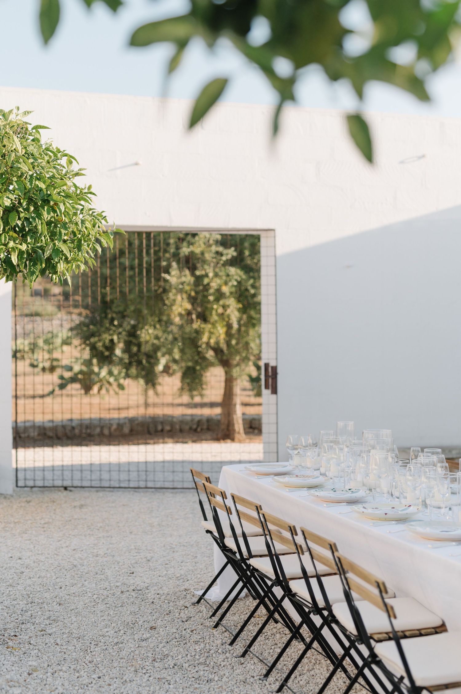 Long wooden tables with black chairs set up for an outdoor event on gravel with trees in the background.