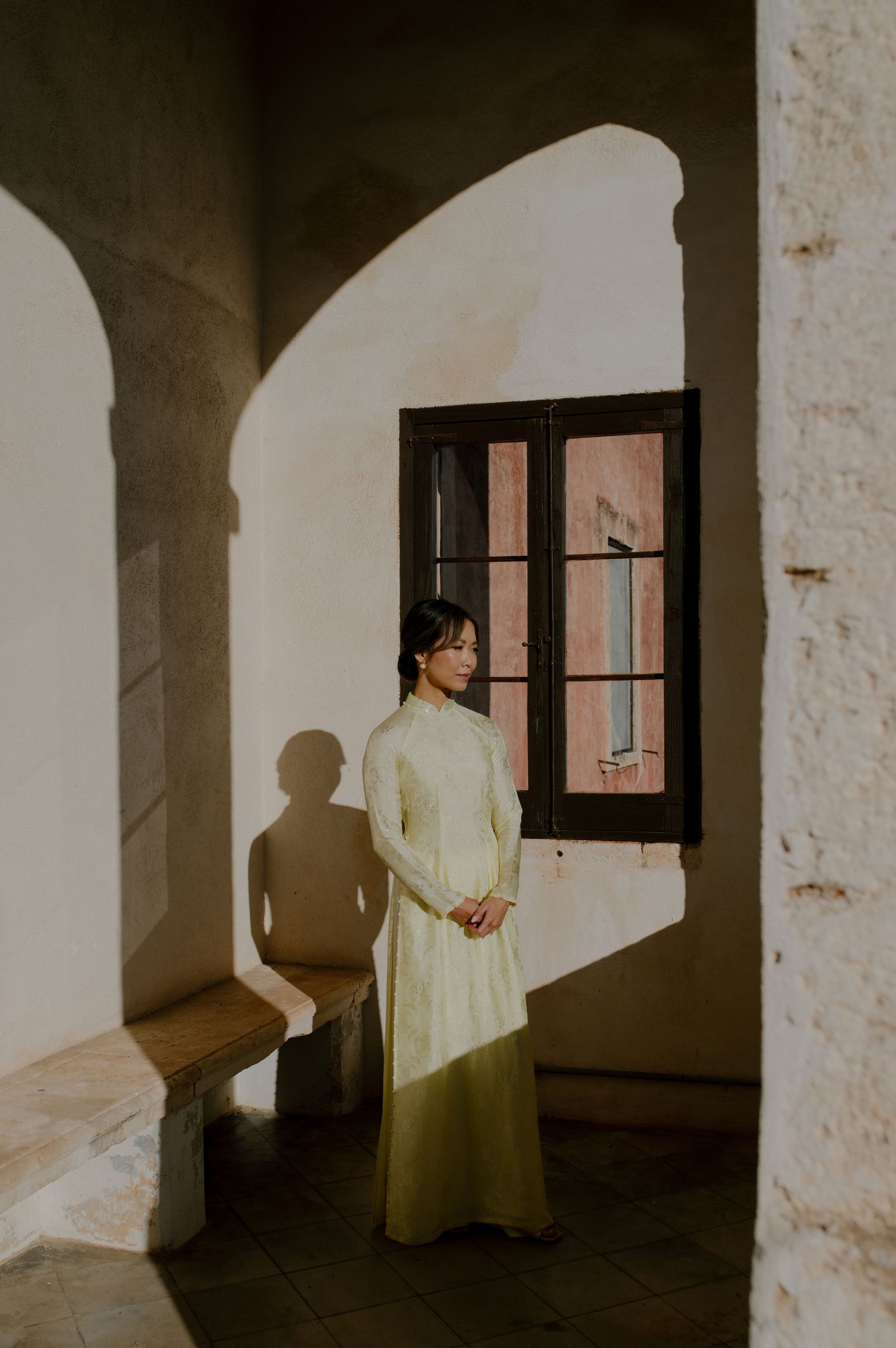A person in a cream-colored dress stands by an arched window, casting dramatic shadows in warm sunlight.