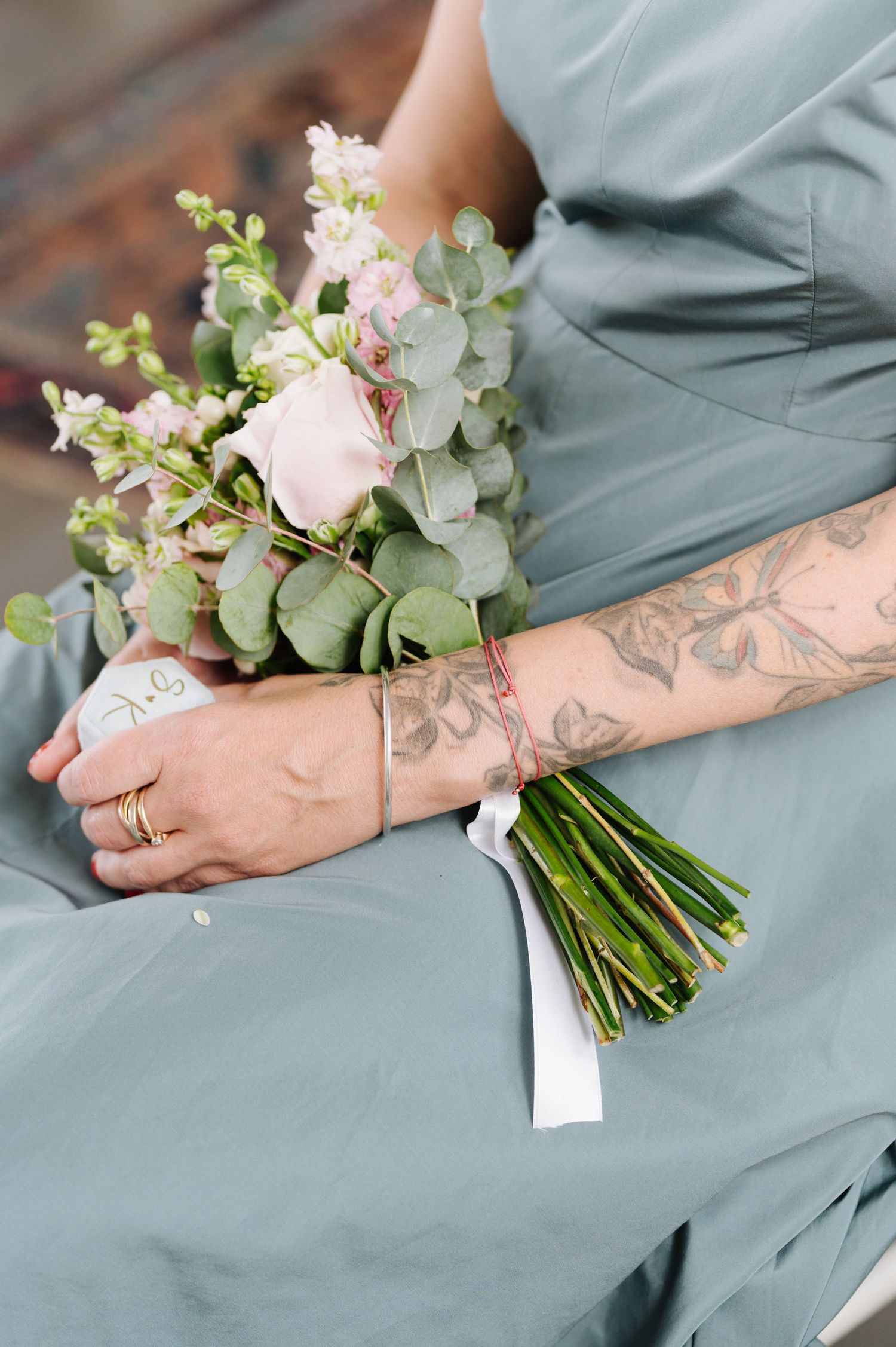 A tattooed arm holds a delicate bouquet of pink roses and eucalyptus against a sage green background.