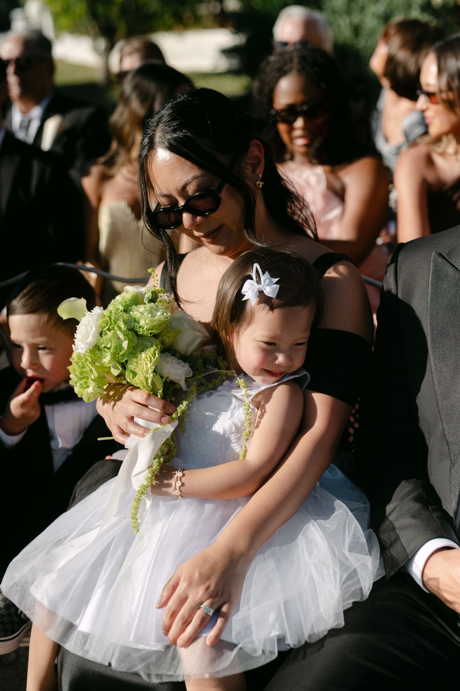 Woman in black dress holds toddler girl in white tutu at outdoor wedding ceremony with green floral bouquet.