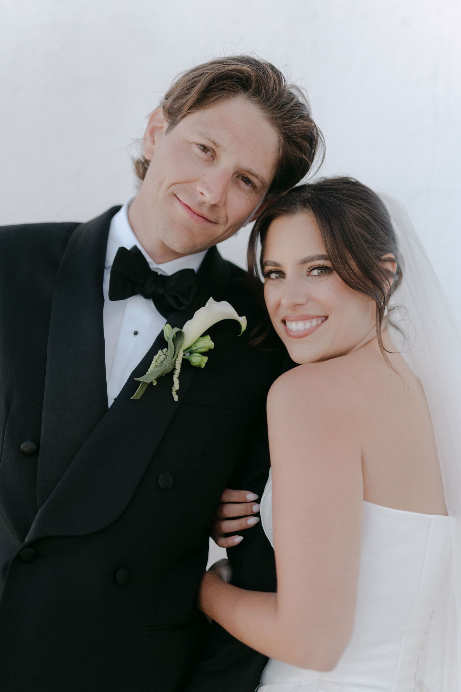 A smiling bride and groom pose together in wedding attire, he in a black tuxedo with boutonniere, she in a white strapless gown.