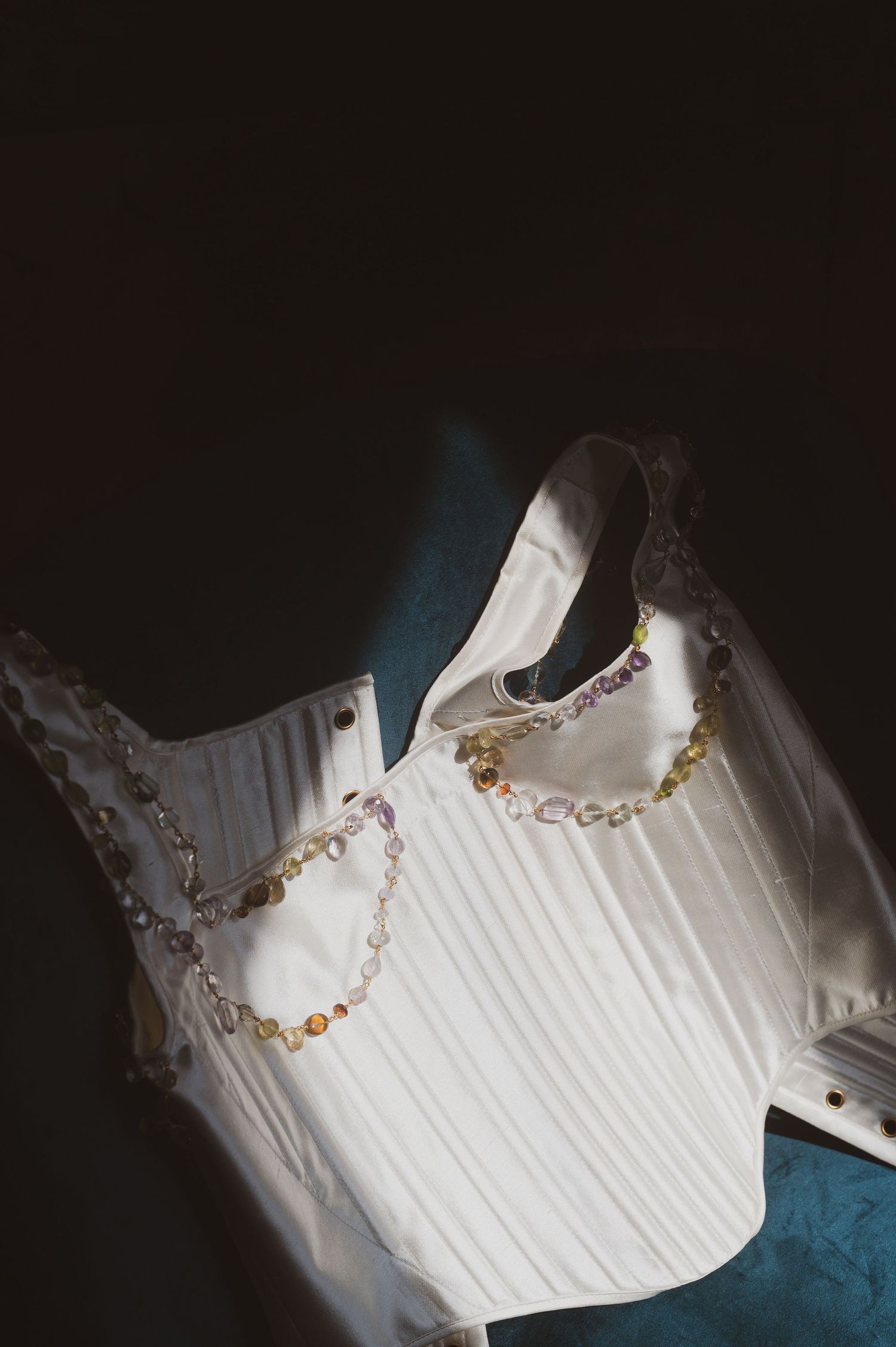 A white corset with delicate gold chain detailing photographed against a dark background with dramatic lighting.