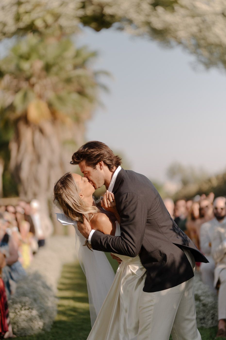 Wedding couple sharing romantic moment under palm trees with guests in background.