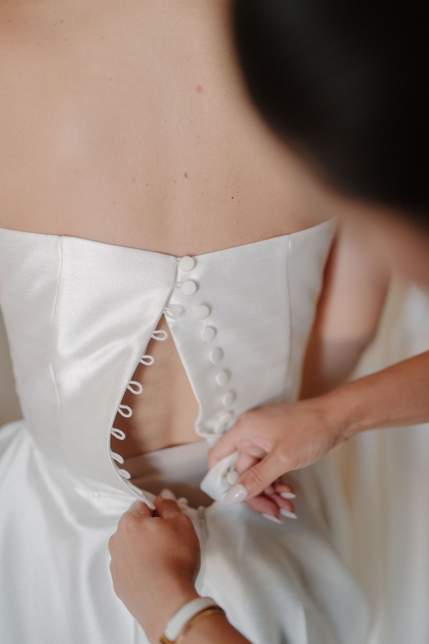 Bridesmaid fastening white satin wedding dress buttons on bride's back during bridal preparation.