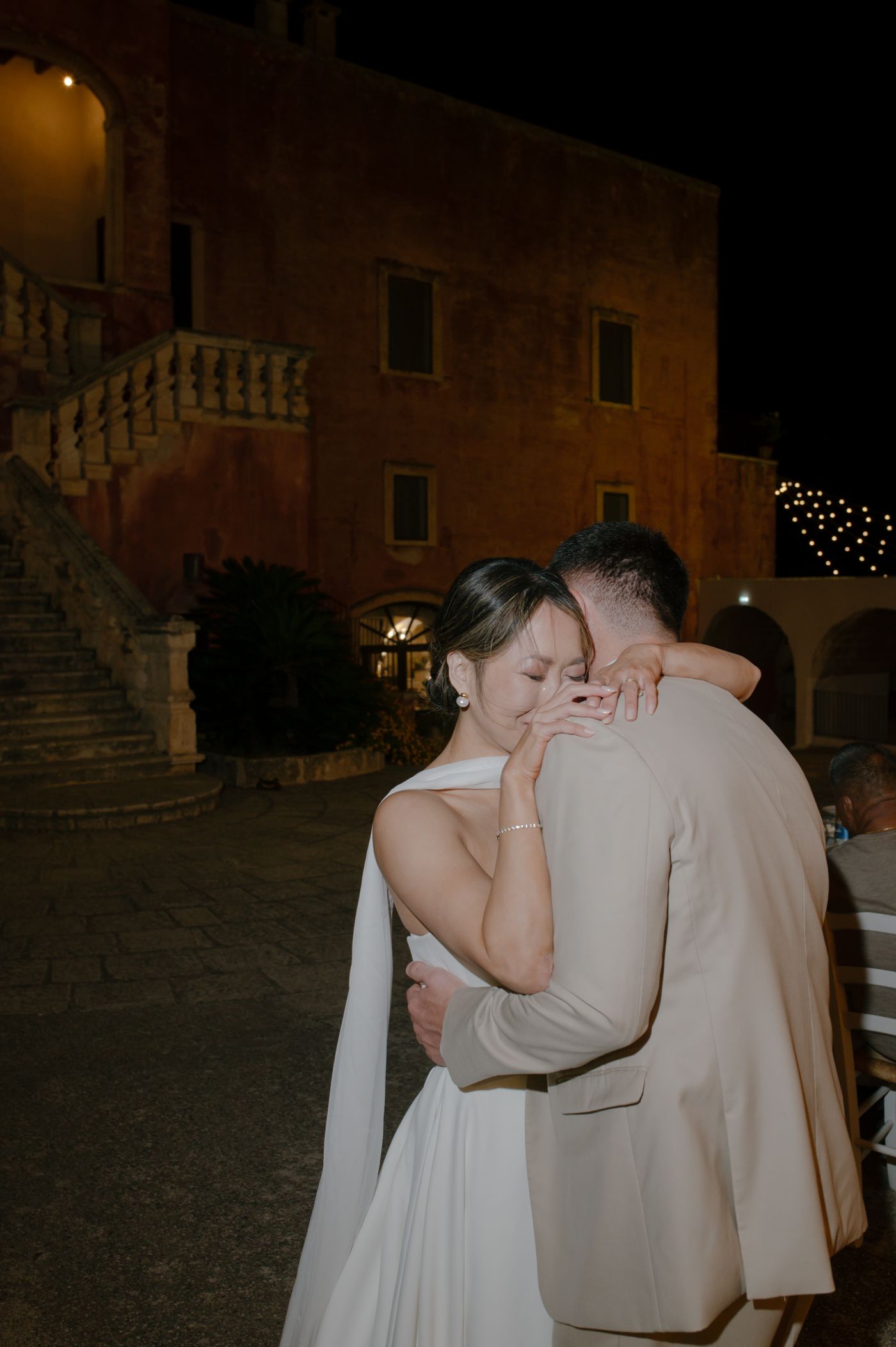 A couple shares an intimate dance moment at night in front of an old stone building with twinkling lights.