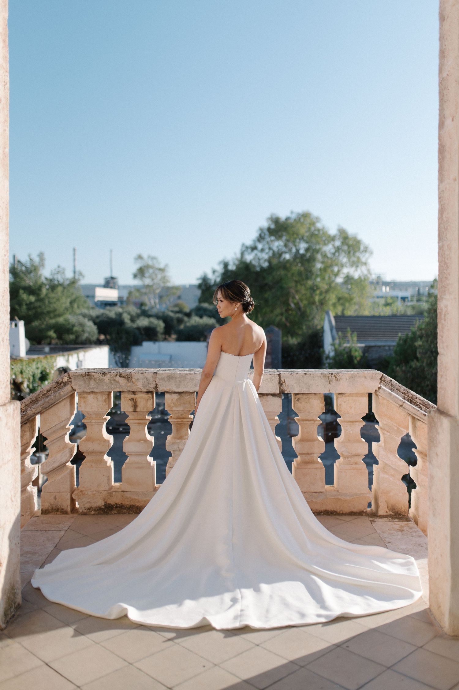 Wedding dress with long flowing train displayed on outdoor stone balcony with ornate balusters against blue sky.