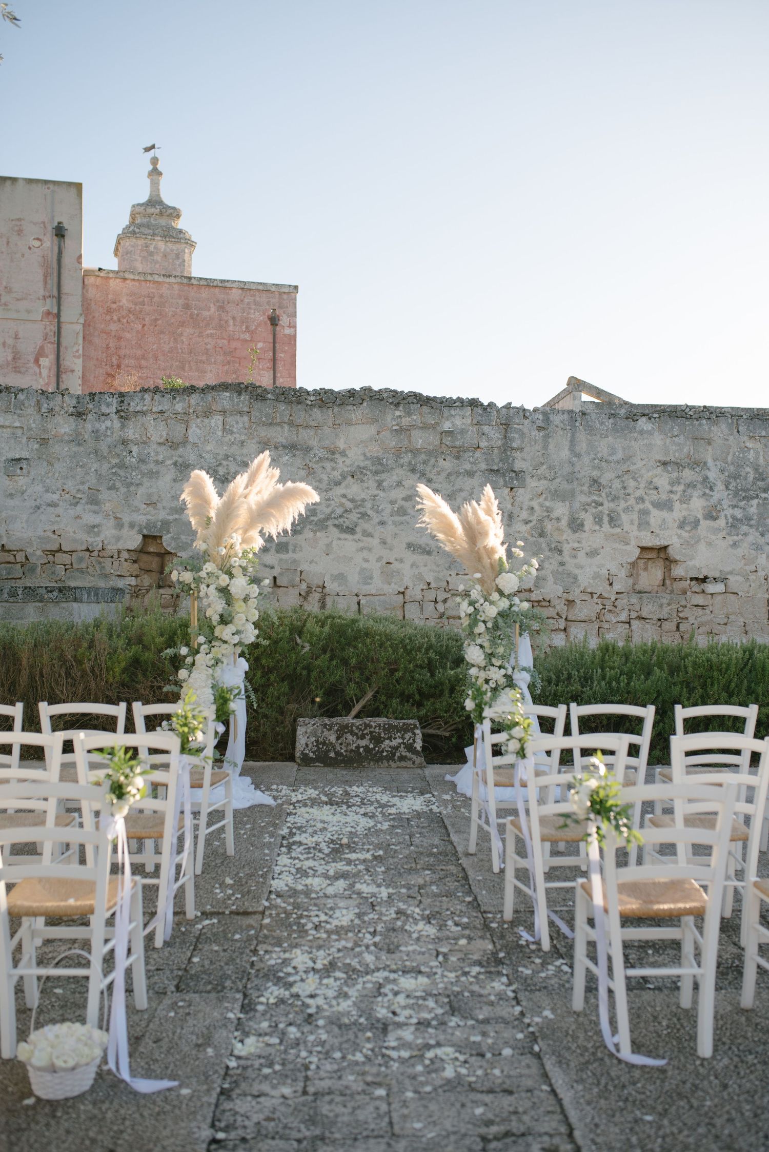 White chairs and decorative arrangements set up for an outdoor wedding ceremony against a rustic stone wall backdrop.