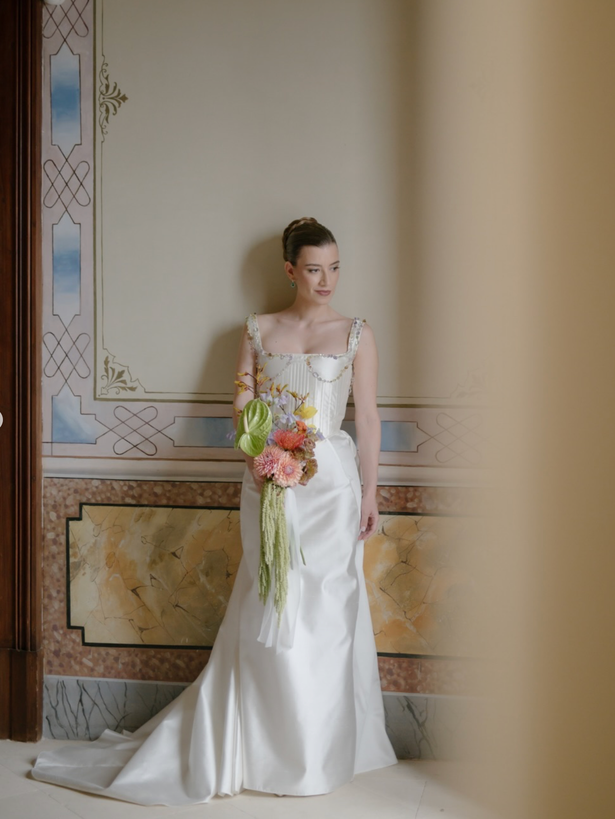 A bride in a classic white wedding gown stands elegantly against an ornate wall with decorative trim and window details.