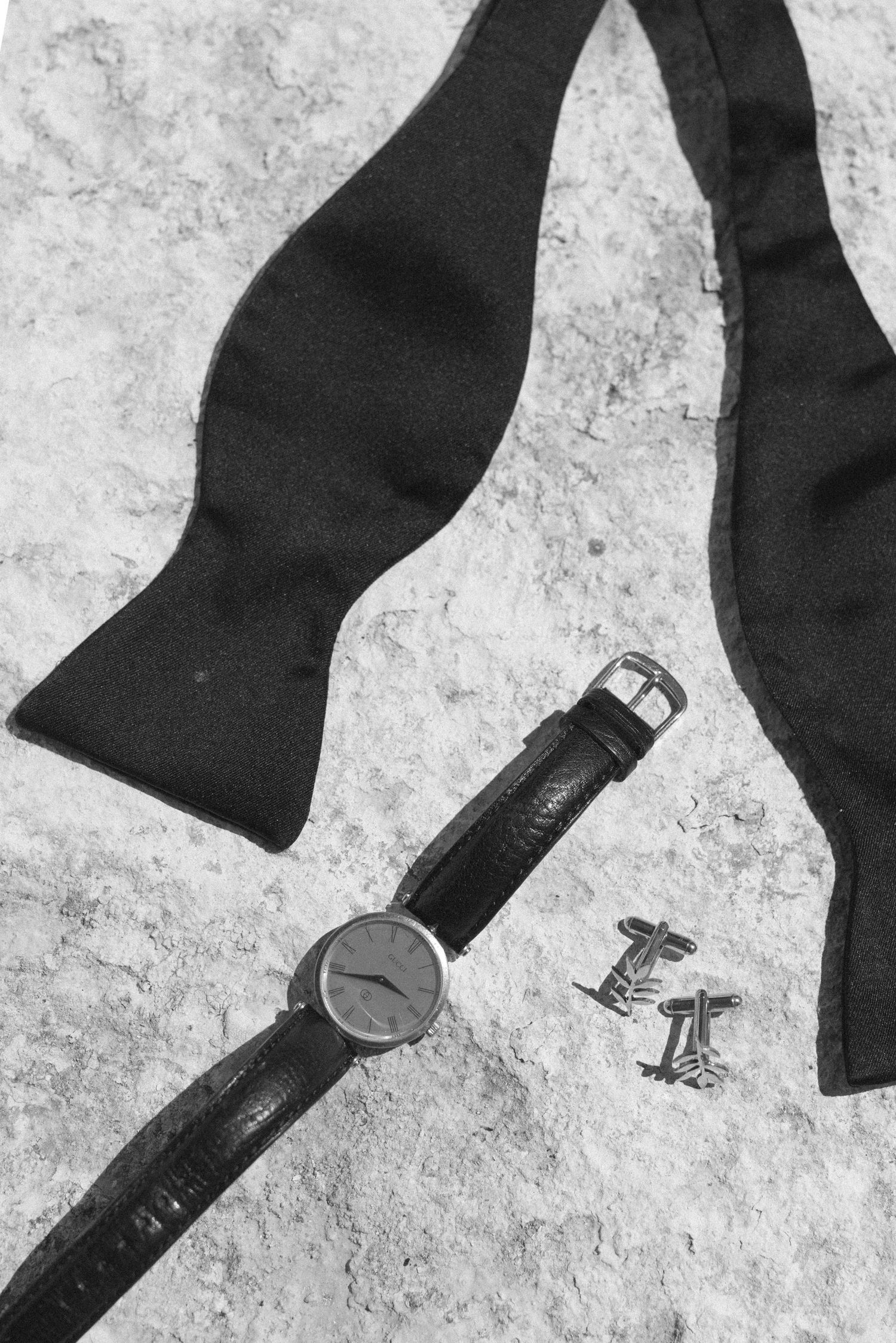 Black bow tie, wristwatch with leather strap, and cufflinks arranged on stone surface in elegant flat lay.