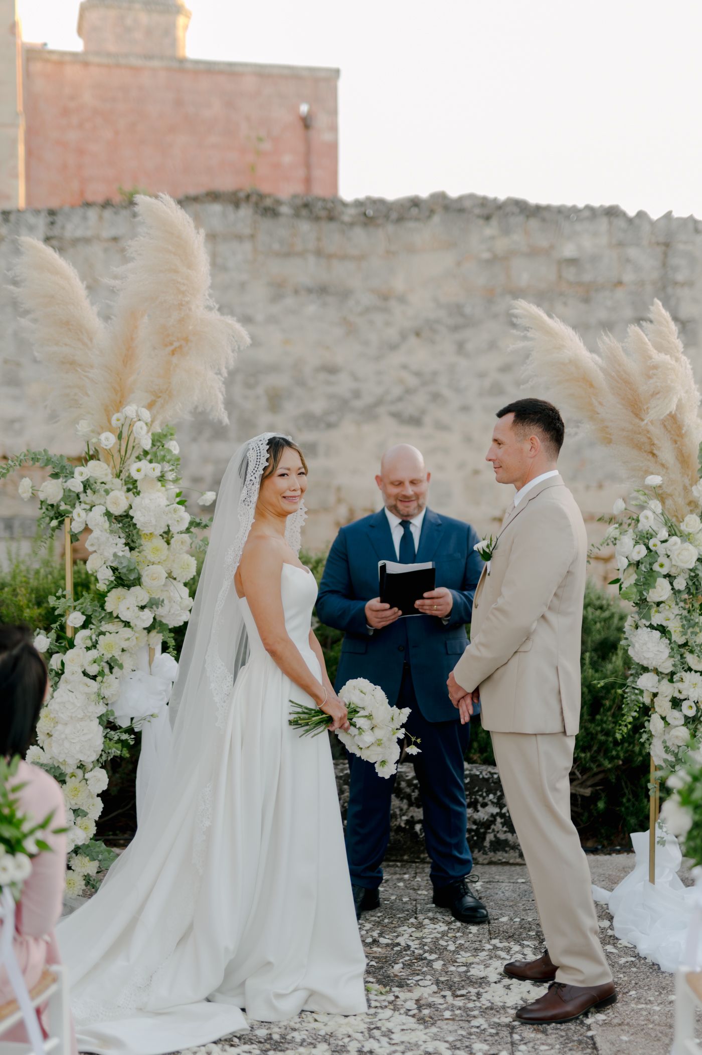 An outdoor wedding ceremony takes place against a stone wall backdrop with white floral arrangements and pampas grass decor.
