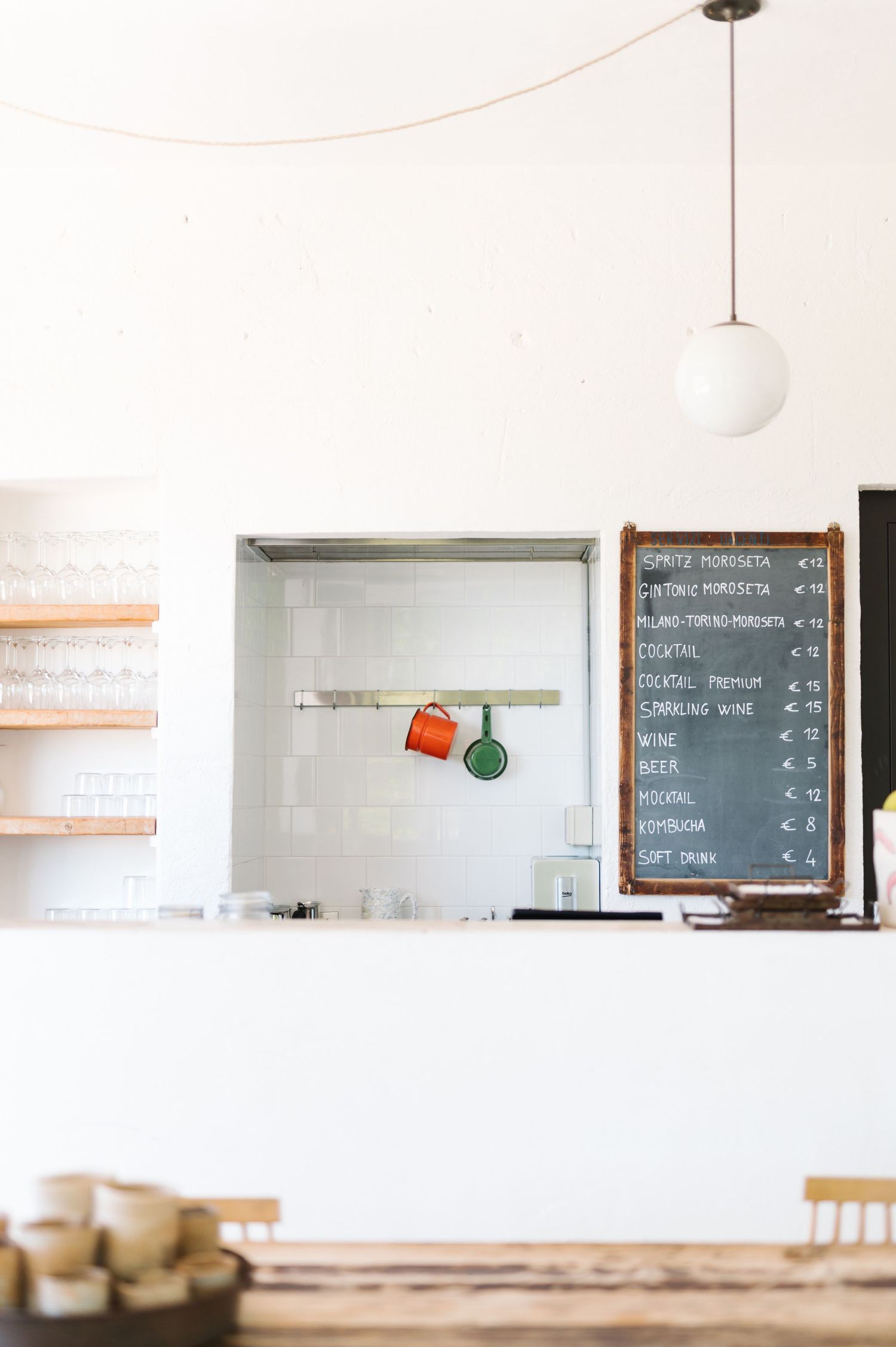 Minimalist cafe interior with white walls, wooden shelves, hanging globe light, and chalkboard menu displaying prices.