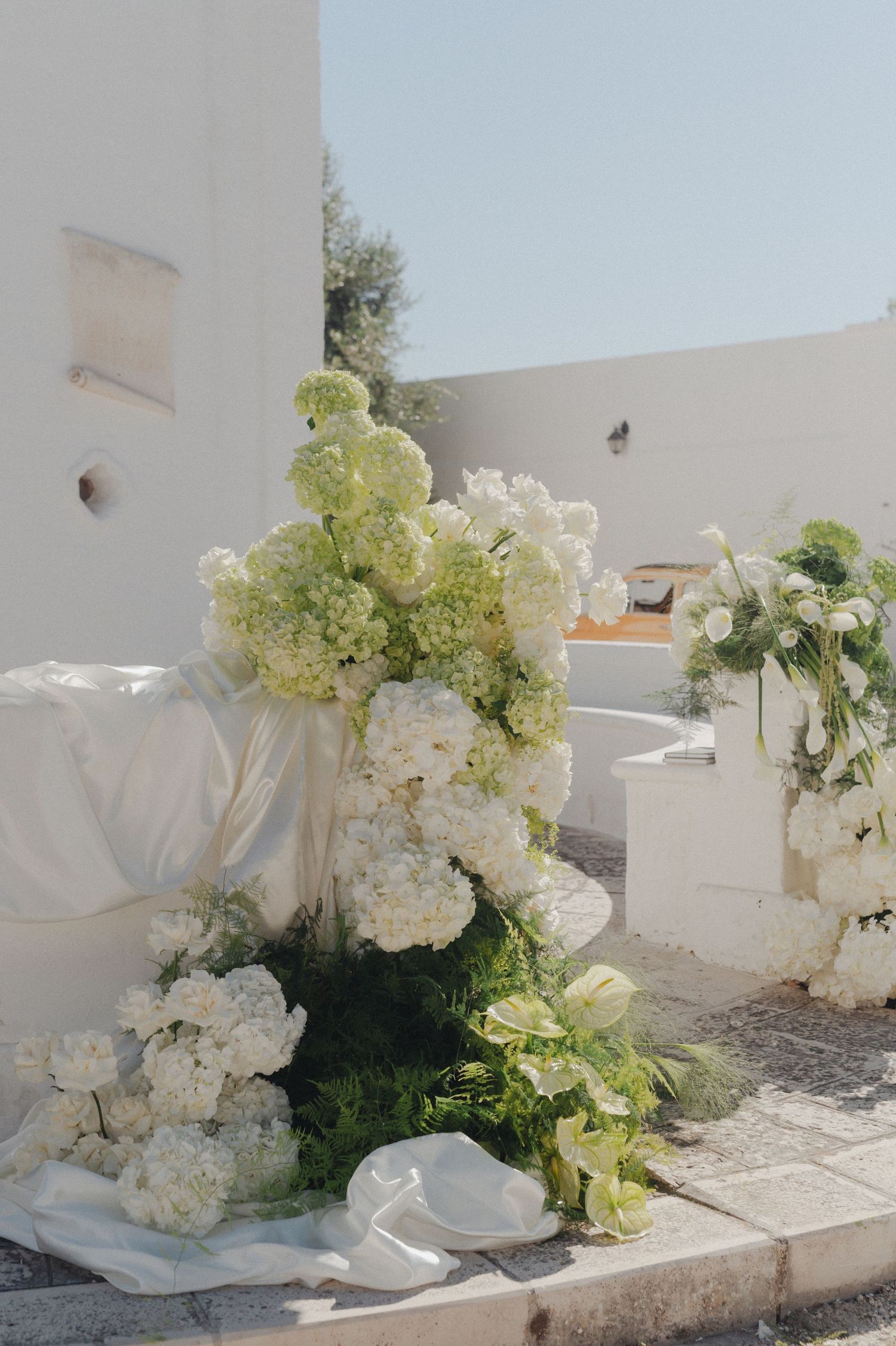 Lush white hydrangea and green floral arrangements adorn a sunlit white stone wedding venue exterior.