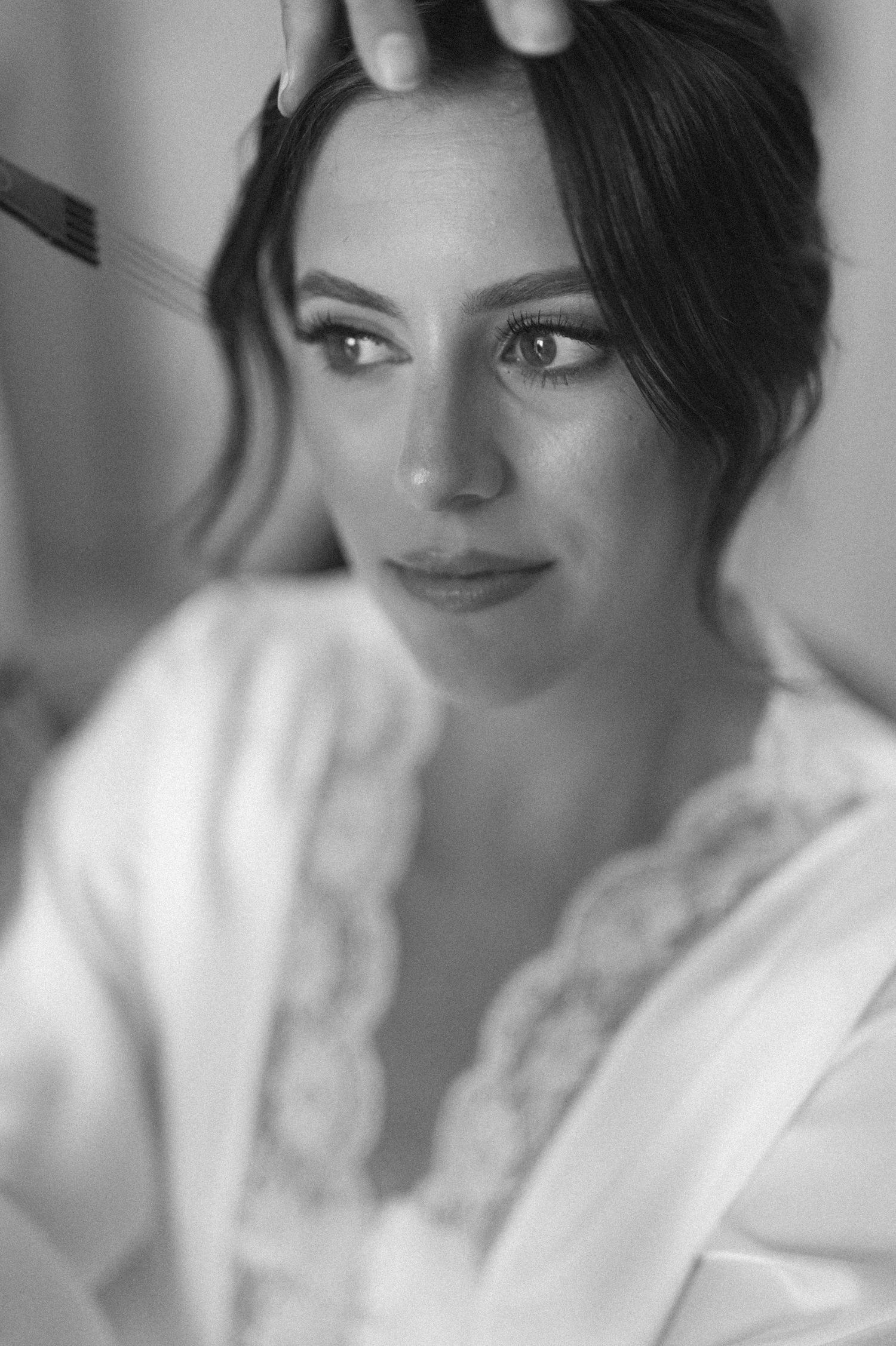 Black and white bridal portrait of a woman in a lace robe getting her hair styled before her wedding.