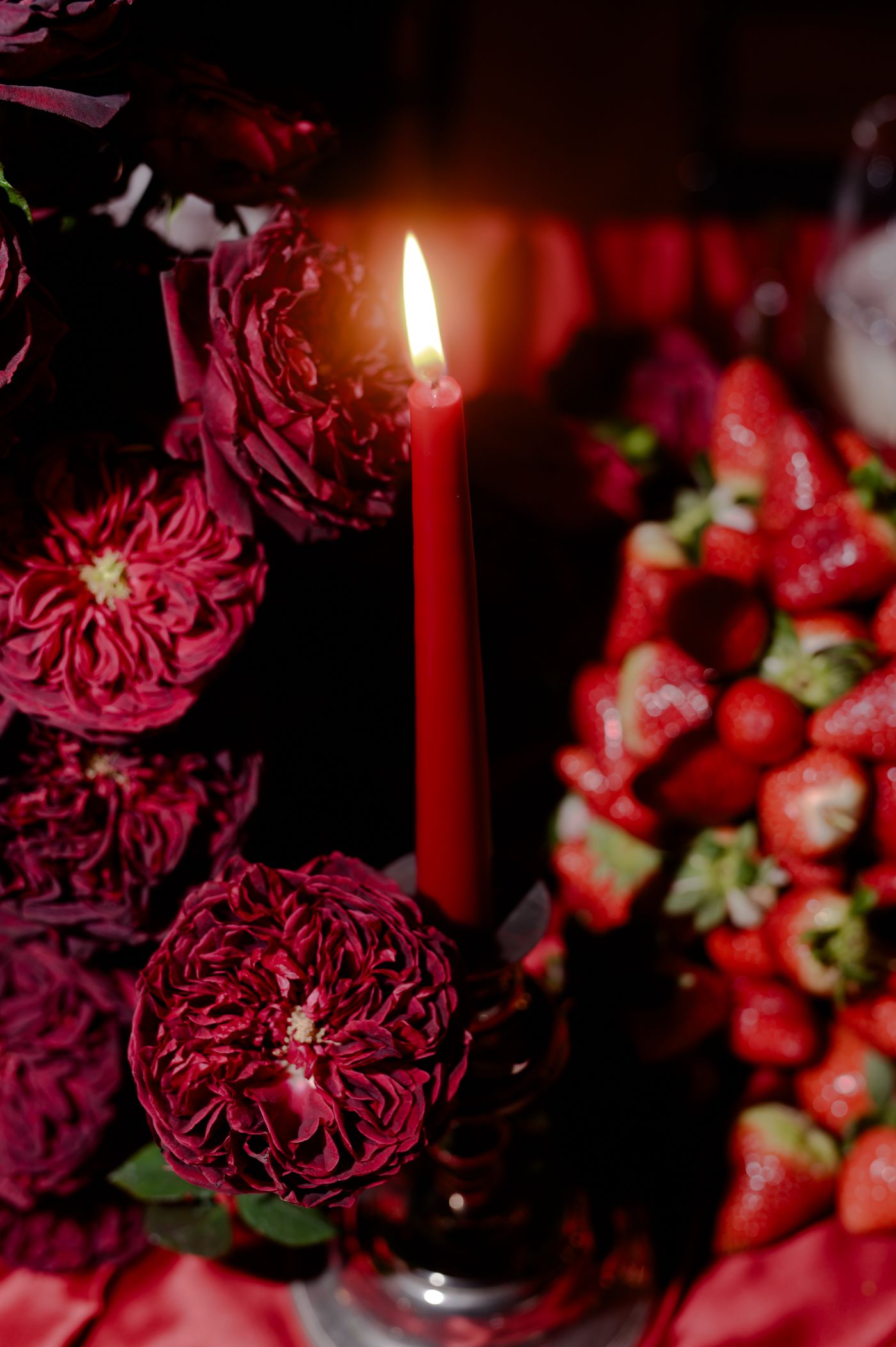 Deep burgundy carnations and red strawberries create a moody composition with glowing red candles in dark lighting.