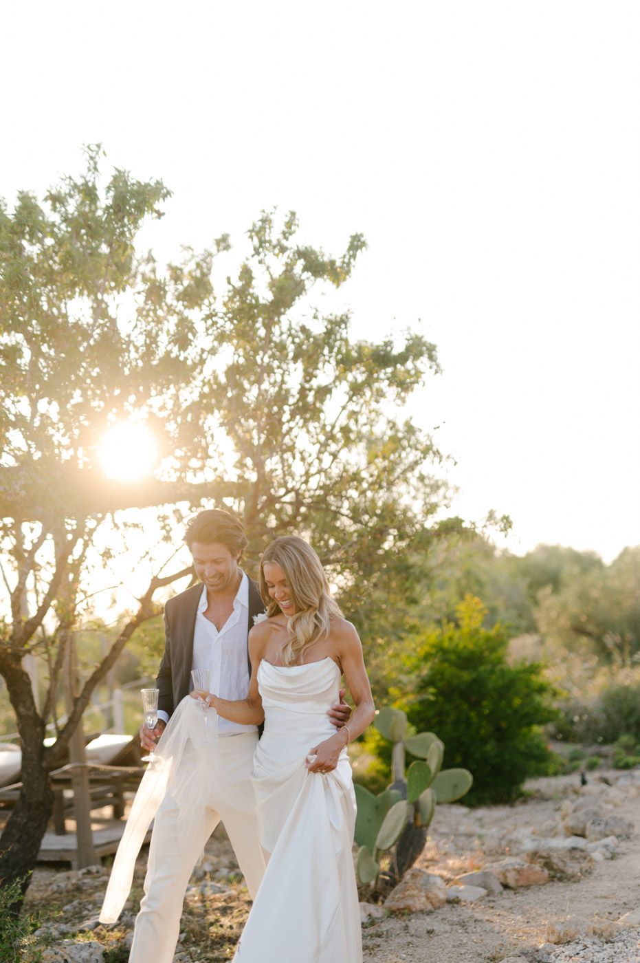 A couple in elegant white attire embraces during golden hour in a desert garden setting with sunlight filtering through trees.
