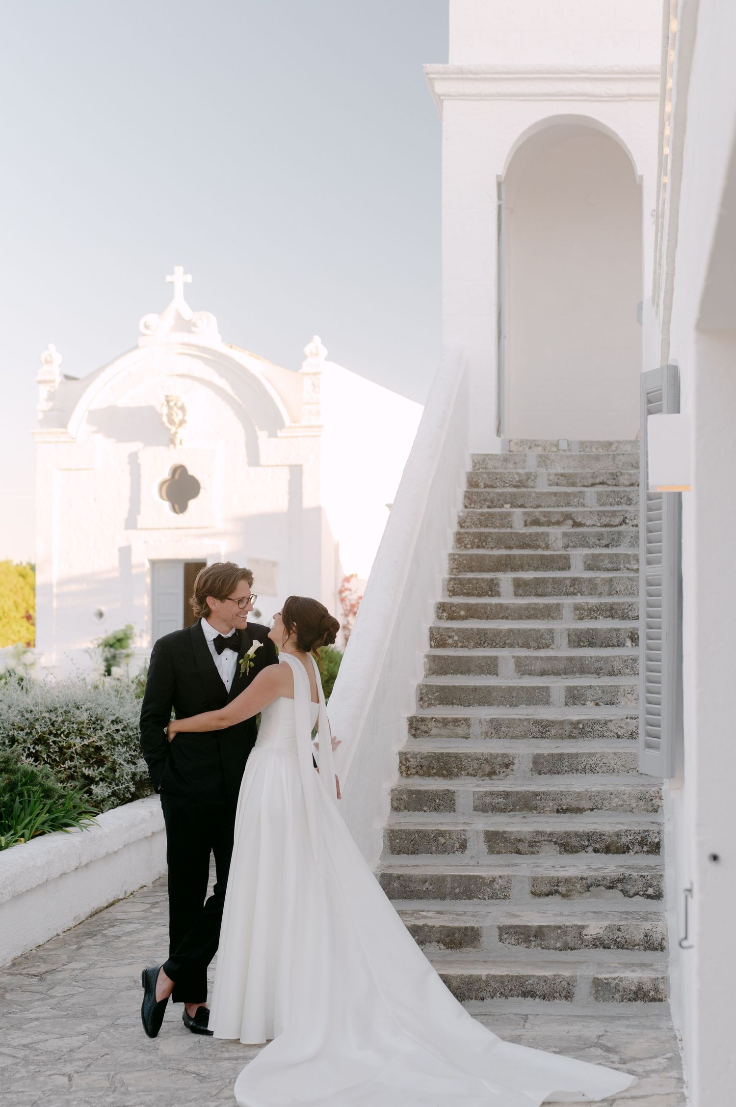 Bride and groom embrace near white chapel stairs in elegant wedding attire, veil flowing, at a Mediterranean venue.