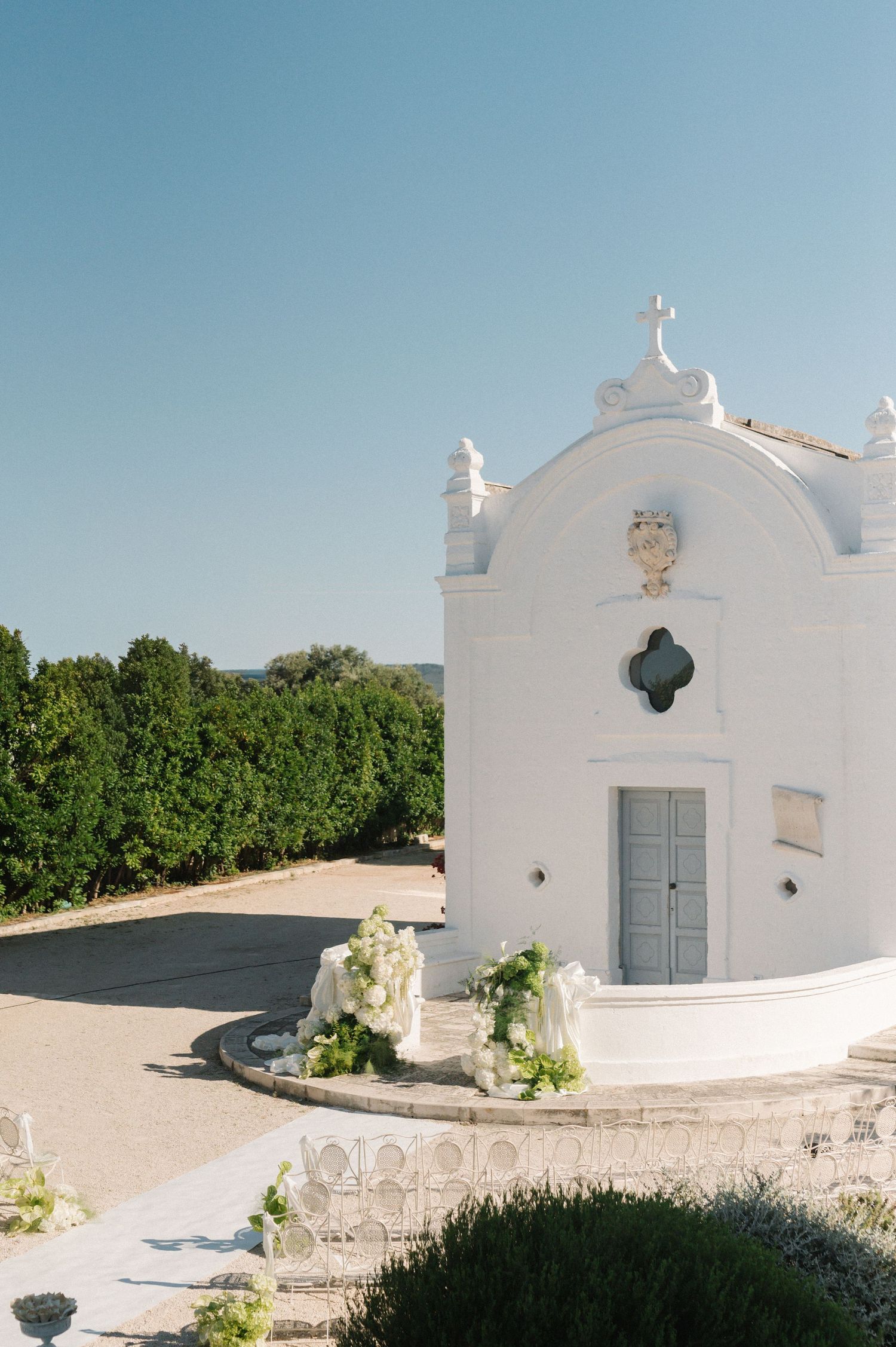 White Mediterranean chapel with floral arrangements and cross atop, surrounded by lush greenery under clear blue sky.