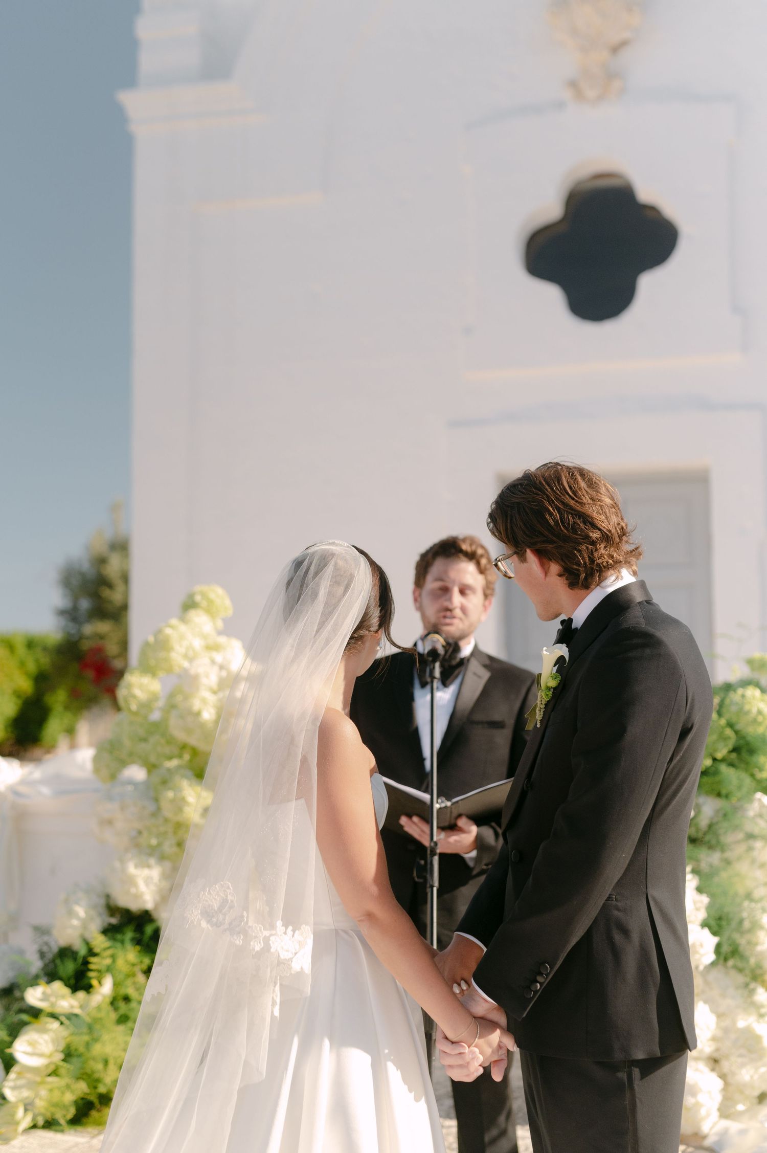 Bride and groom exchange vows outdoors before a white chapel with floral arrangements and an officiant.