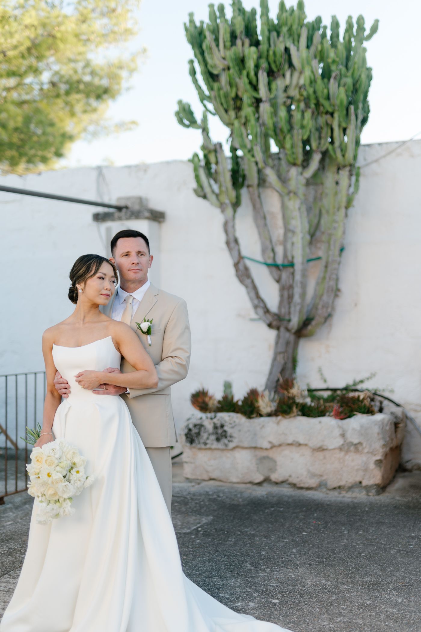 A couple poses for a wedding photo in front of a tall cactus plant and white wall on their special day.