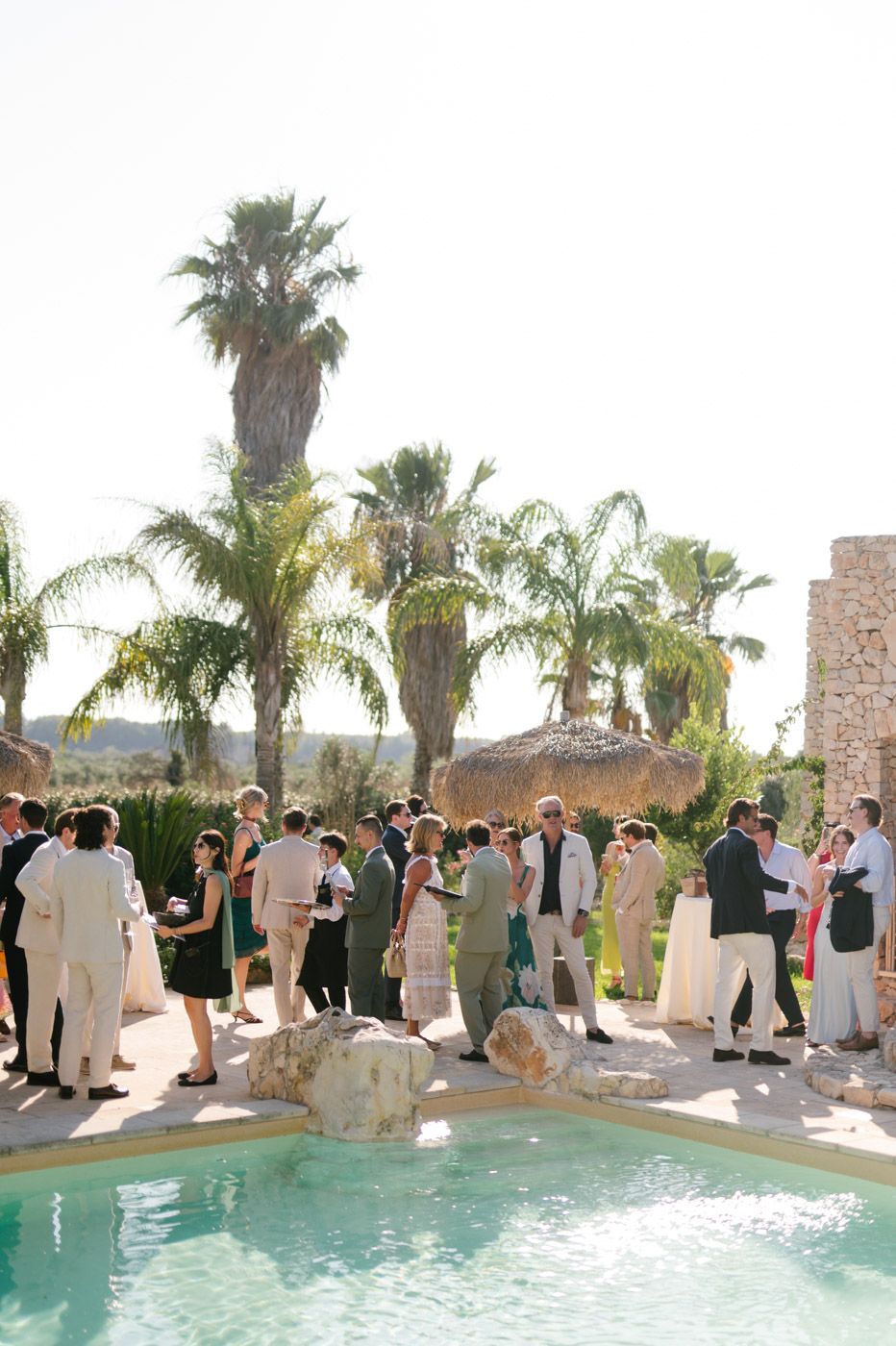 Wedding guests mingling around a resort pool with palm trees.
