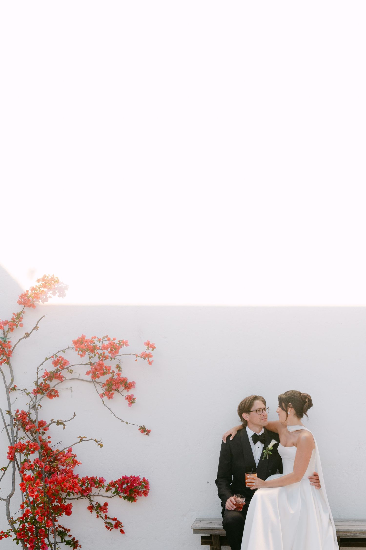 Bride and groom share a romantic moment beside red bougainvillea against a white wall.