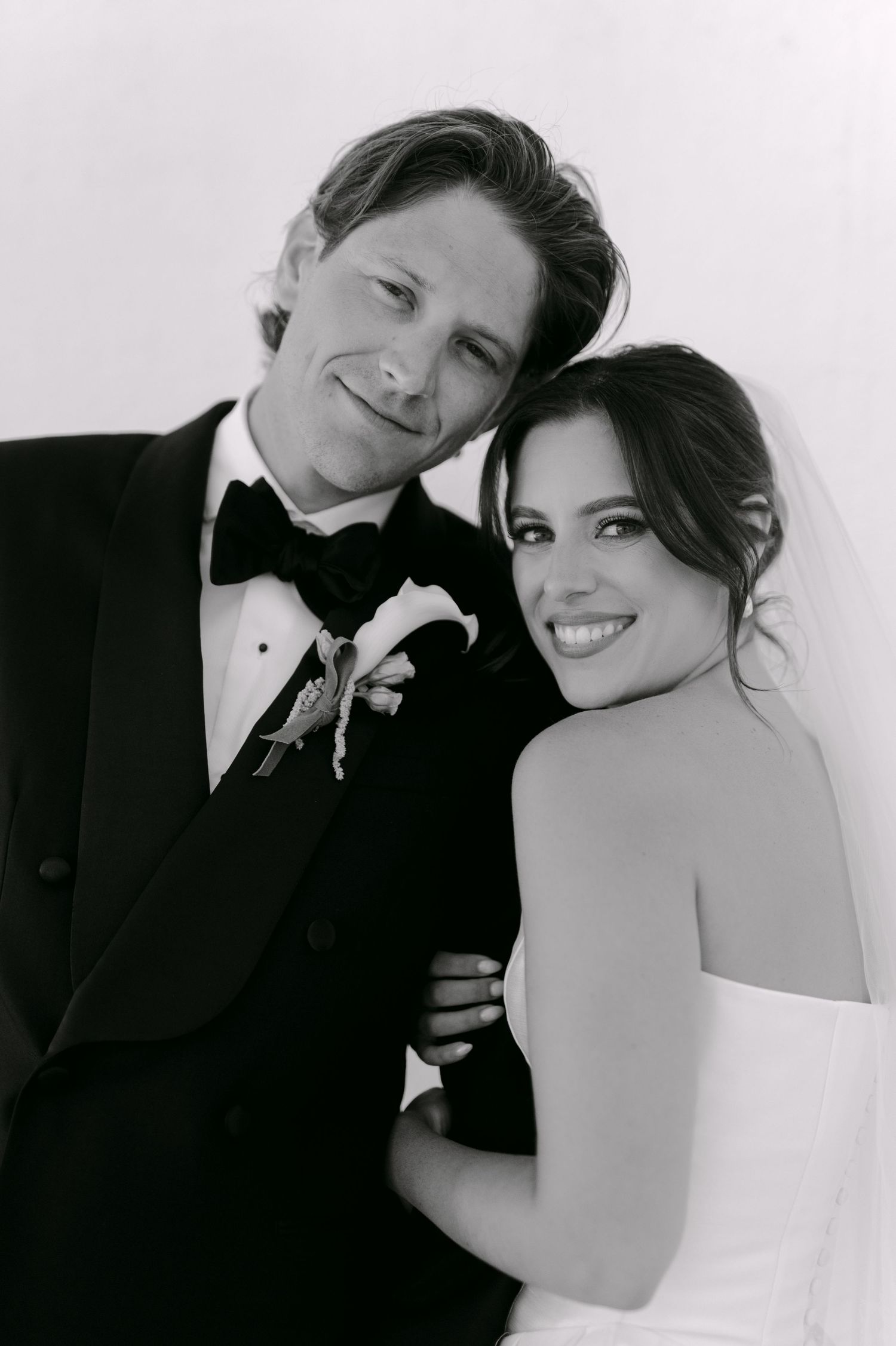 Black and white wedding portrait of smiling bride in veil and groom in tuxedo with boutonniere embracing together.