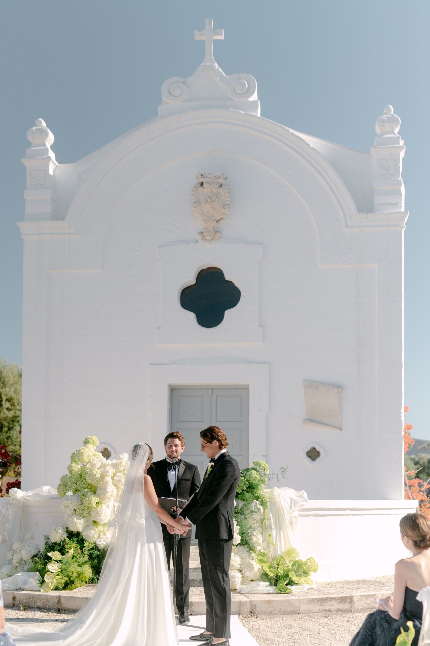 Couple exchanges vows outdoors before a white Mediterranean chapel adorned with floral arrangements on a sunny wedding day.