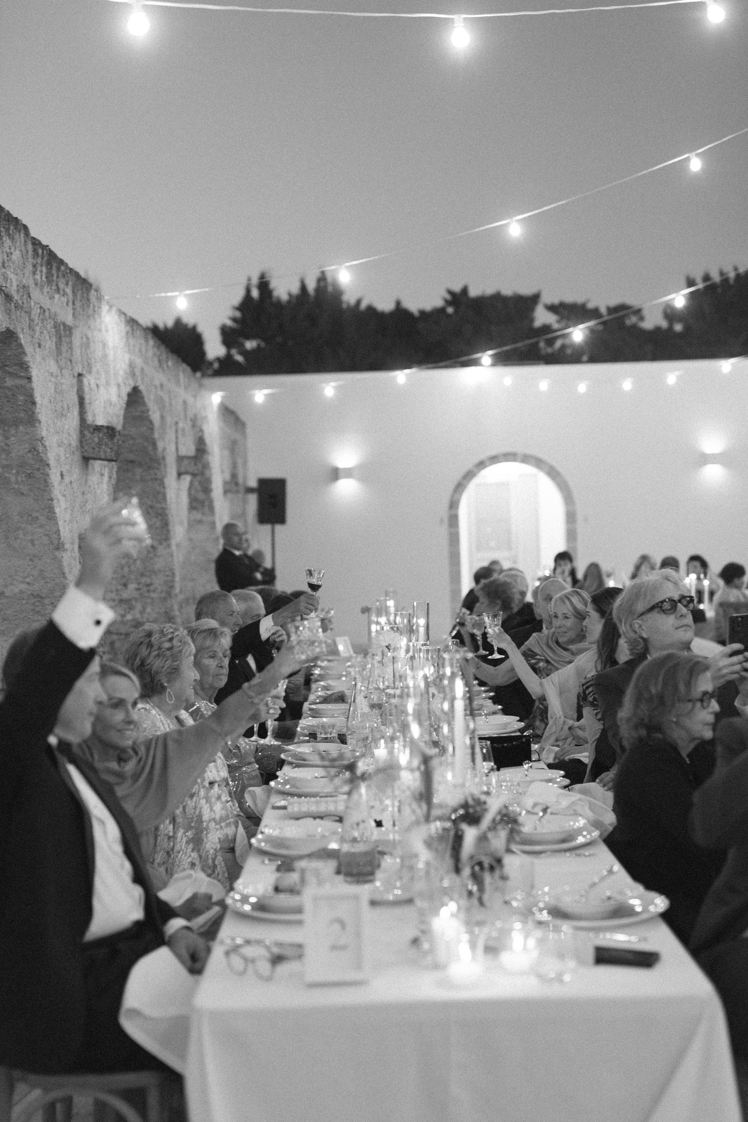 Black and white photo of elegant outdoor dinner party with string lights, long decorated table, and guests raising glasses.